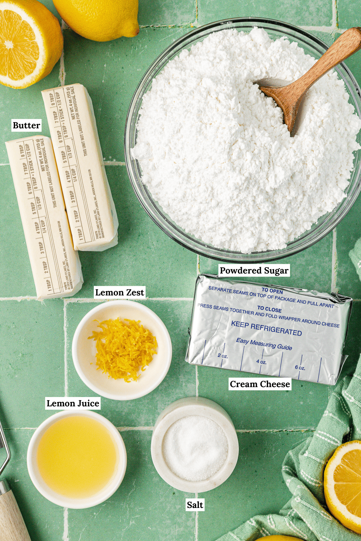 over head view of the ingredients for lemon cream cheese frosting including fresh lemons, powdered sugar, two sticks of nutter, lemon zest, a block of cream cheese, lemon juice, and salt