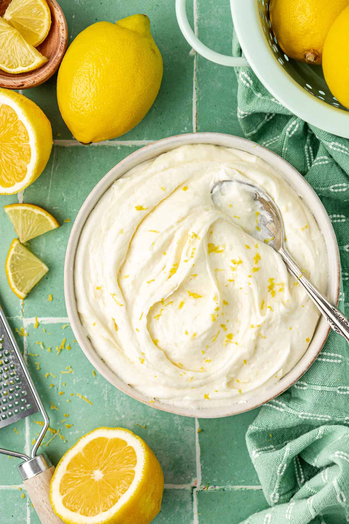 over head view of a bowl of lemon cream cheese frosting with a spoon in it, topped with lemon zest, surrounded by fresh lemons, lemon slices and wedges, a grater and lemon zest sprinkled around