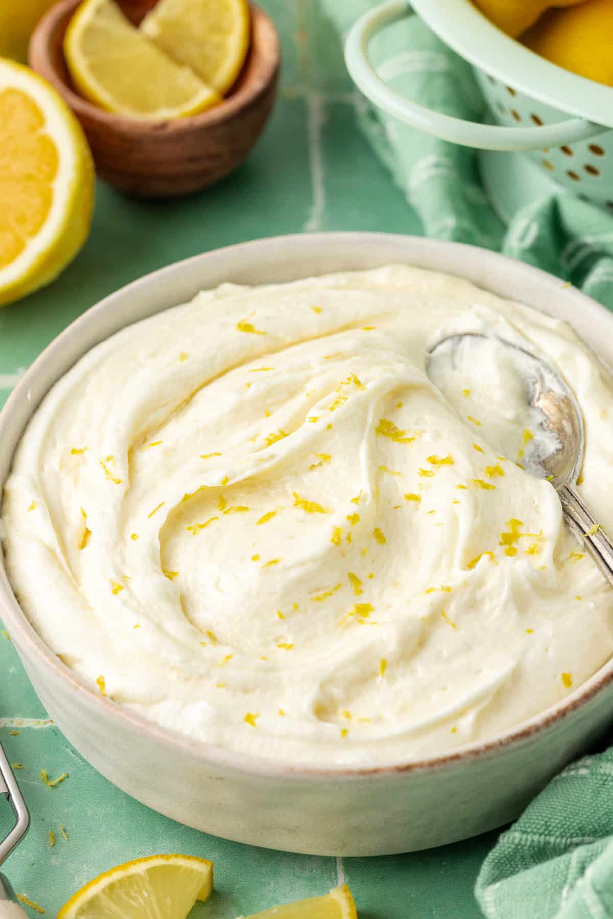 a white bowl of lemon cream cheese frosting with a spoon, topped with lemon zest and surrounded by fresh lemon slices