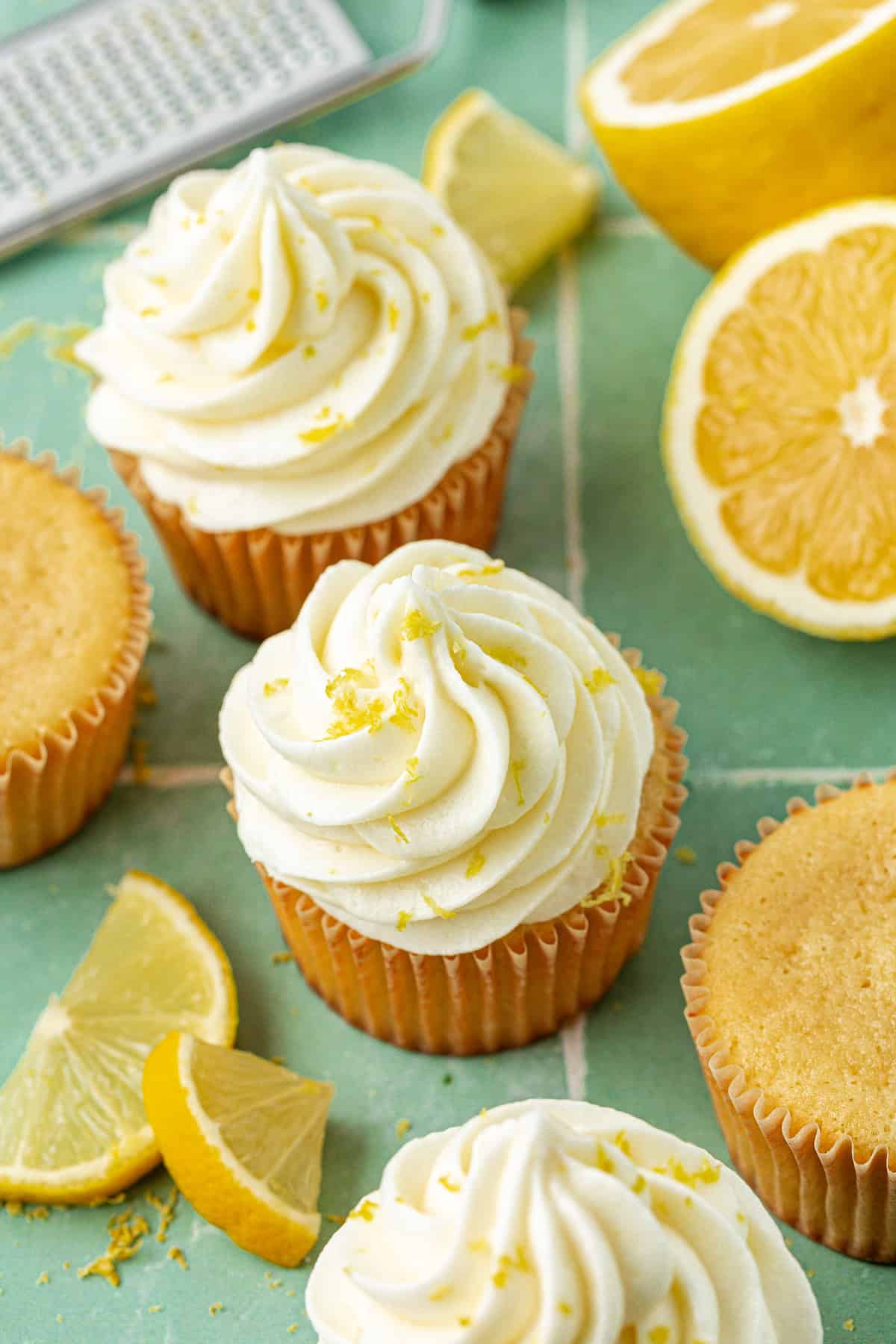 cupcakes topped with lemon cream cheese frosting and sprinkled with lemon zest surrounded by a grater, sliced lemons, and more zest sprinkled around