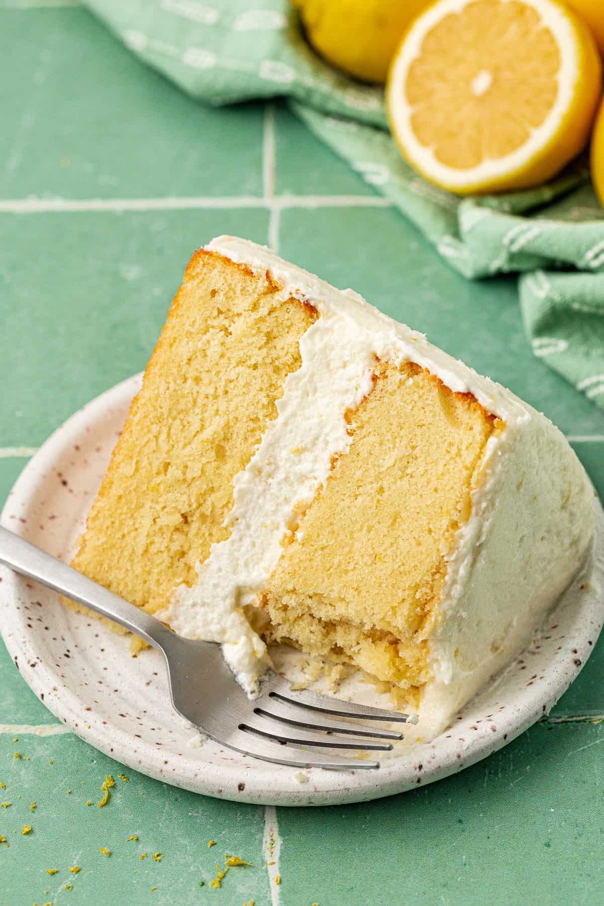 a two layer slice of cake with white frosting on a small plate with a fork and lemons in the background