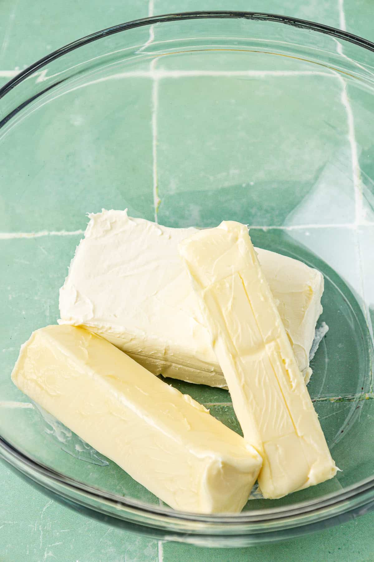 two sticks of butter and a block of cream cheese in a clear glass bowl