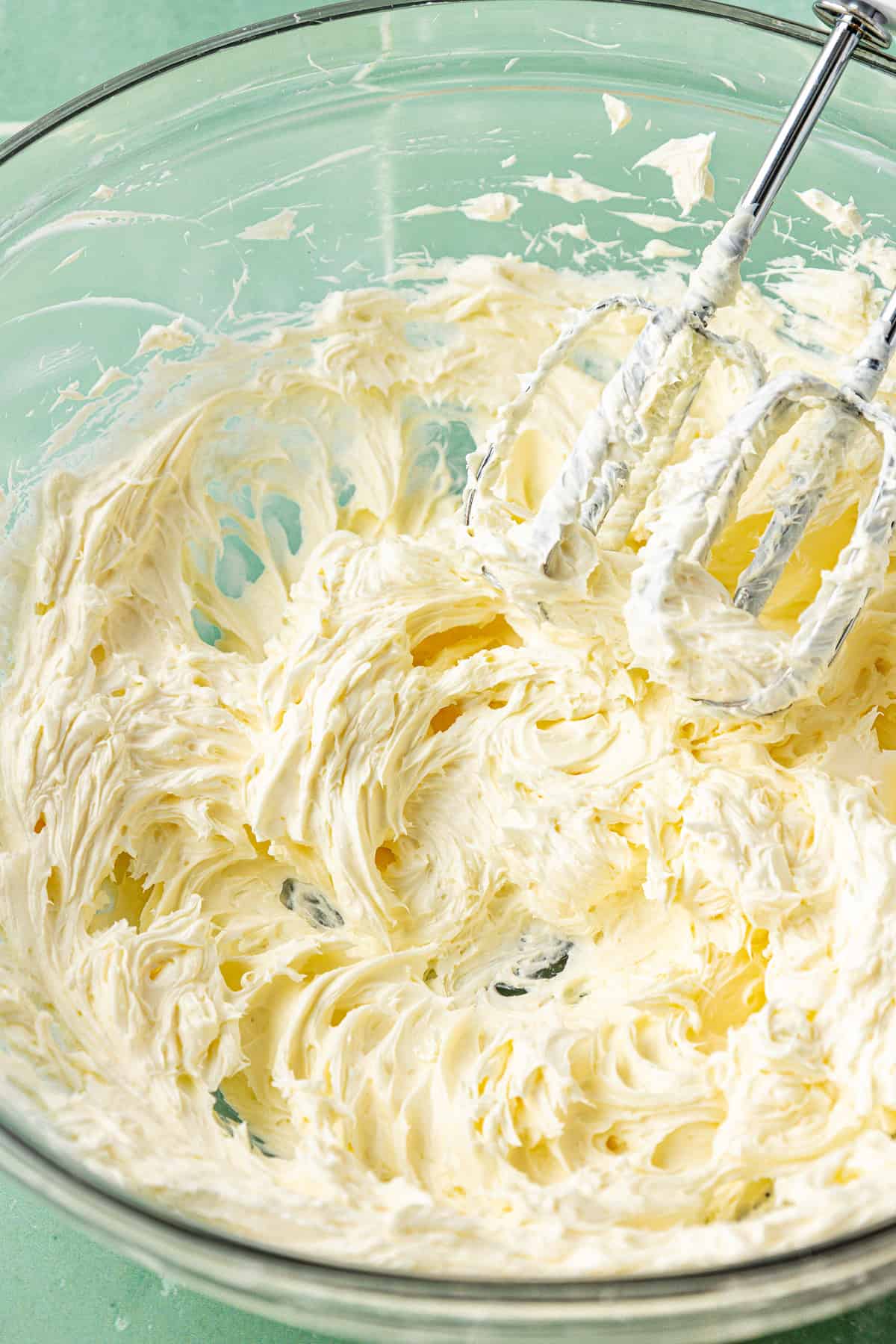 cream cheese and butter creamed together in a large clear glass bowl with an electric mixer leaning in the mixture