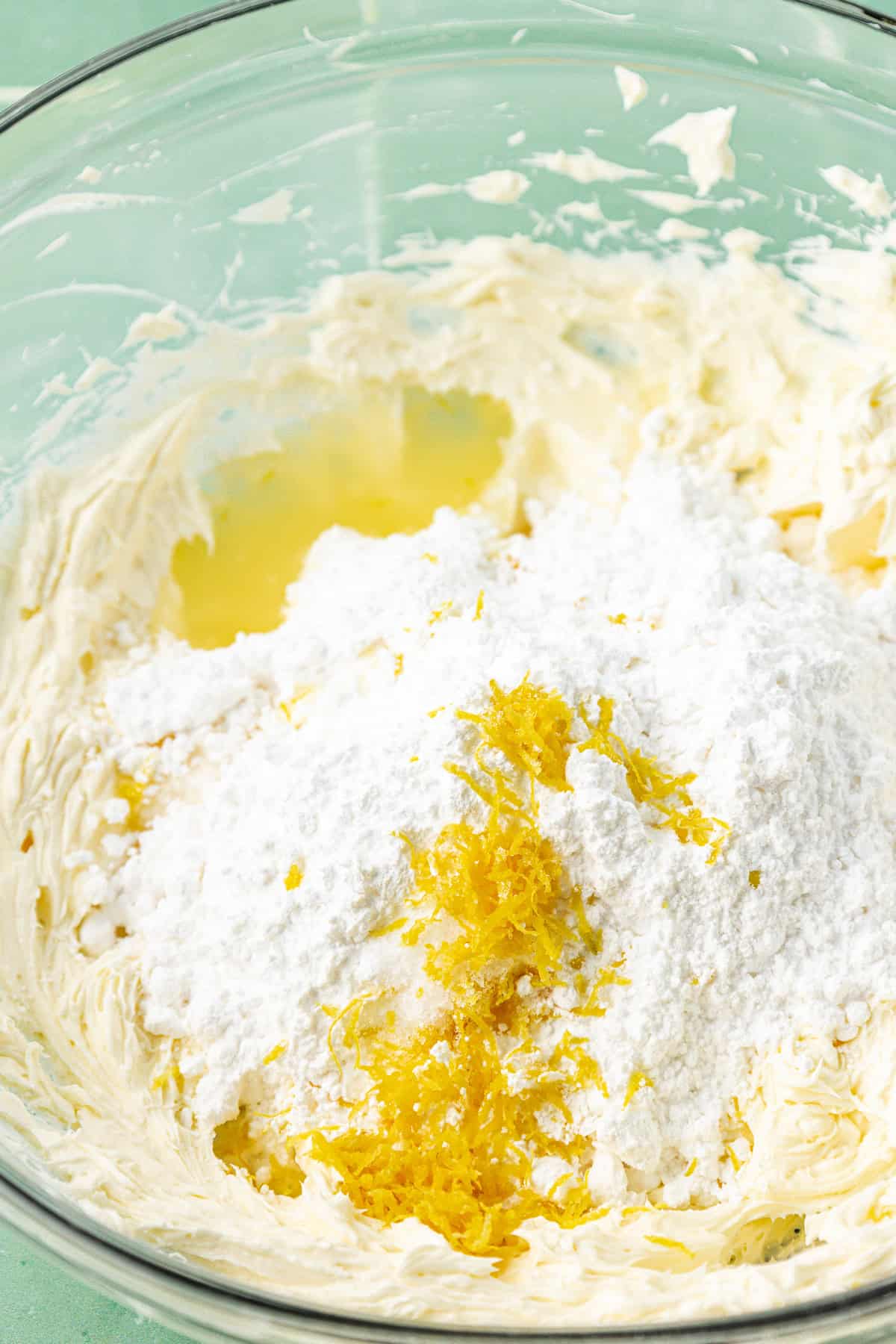 a clear glass bowl with the ingredients for lemon cream cheese frosting, not yet combined, including cream cheese, powdered sugar, and lemon zest