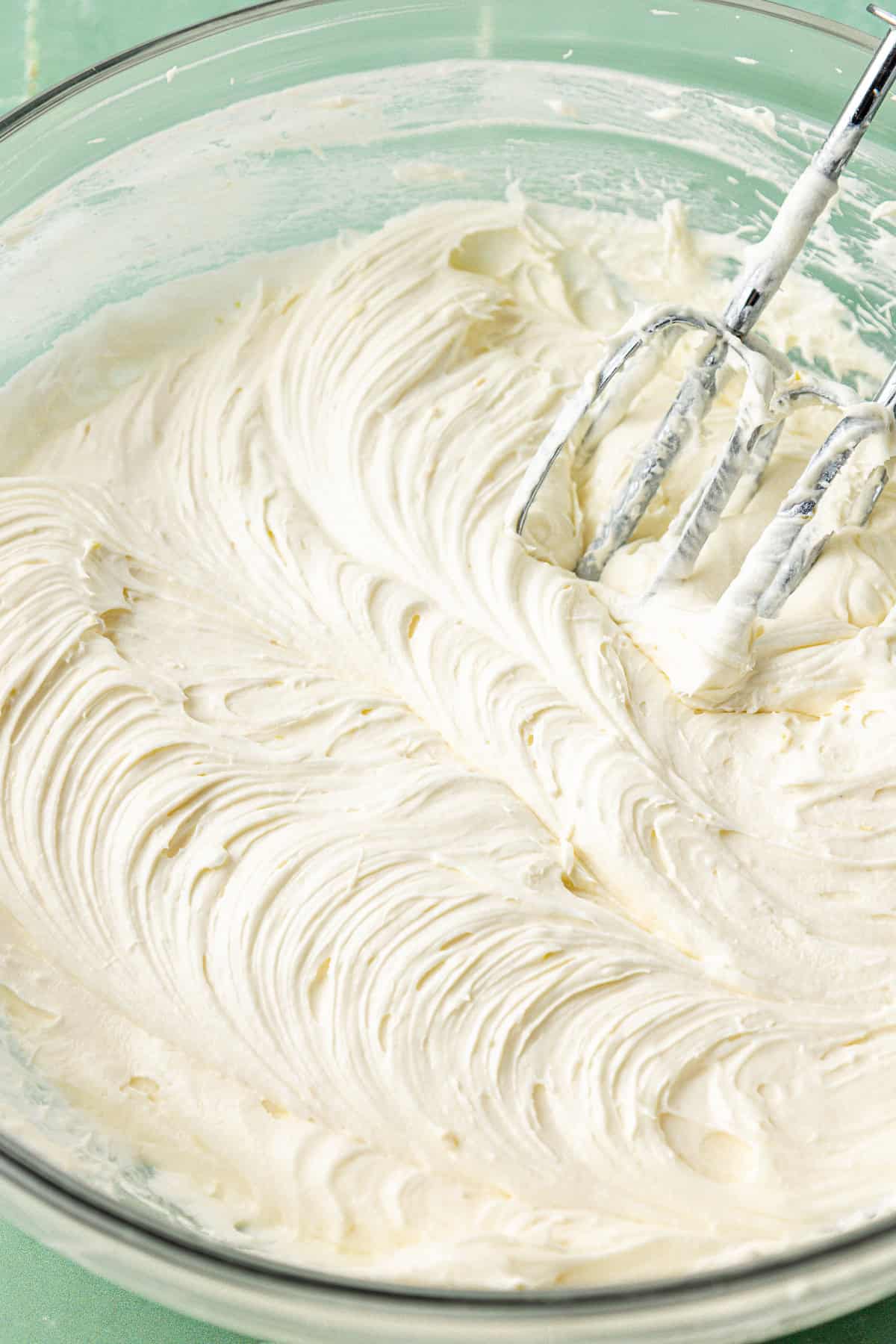 frosting mixed in a large clear glass bowl with an electric mixer leaning in the mixture