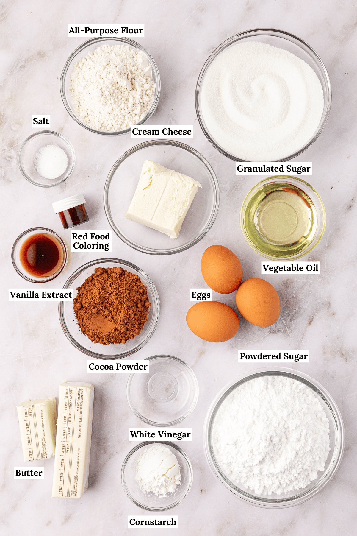 overhead view of the ingredients for red velvet brownies including all-purpose flour, granulated sugar, salt, cream cheese, red food coloring, vanilla extract, cocoa powder, eggs, vegetable oil, butter. white vinegar, cornstarch and powdered sugar