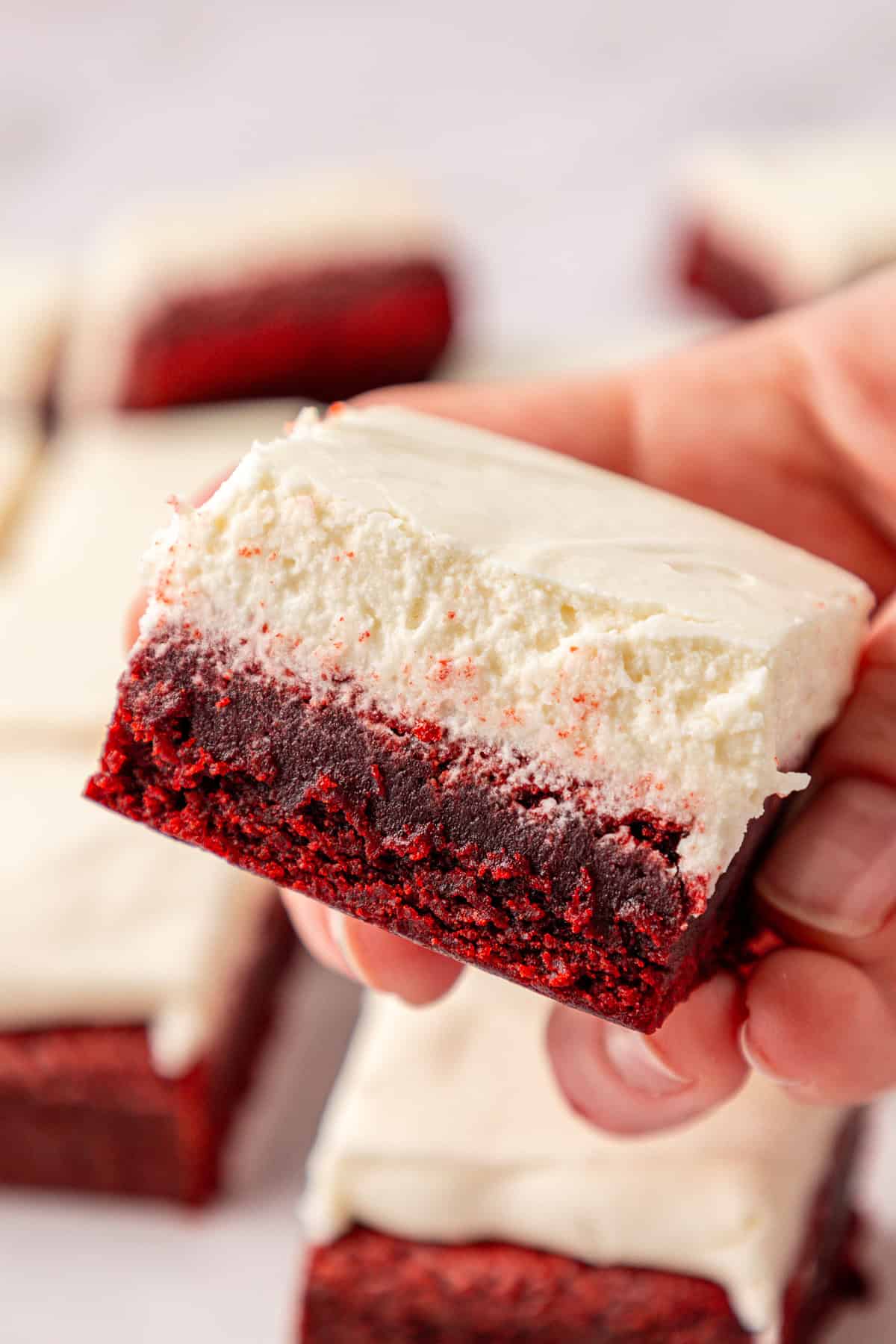 close of up a red velvet brownie topped with cream cheese frosting, held in a hand above more brownies in the background