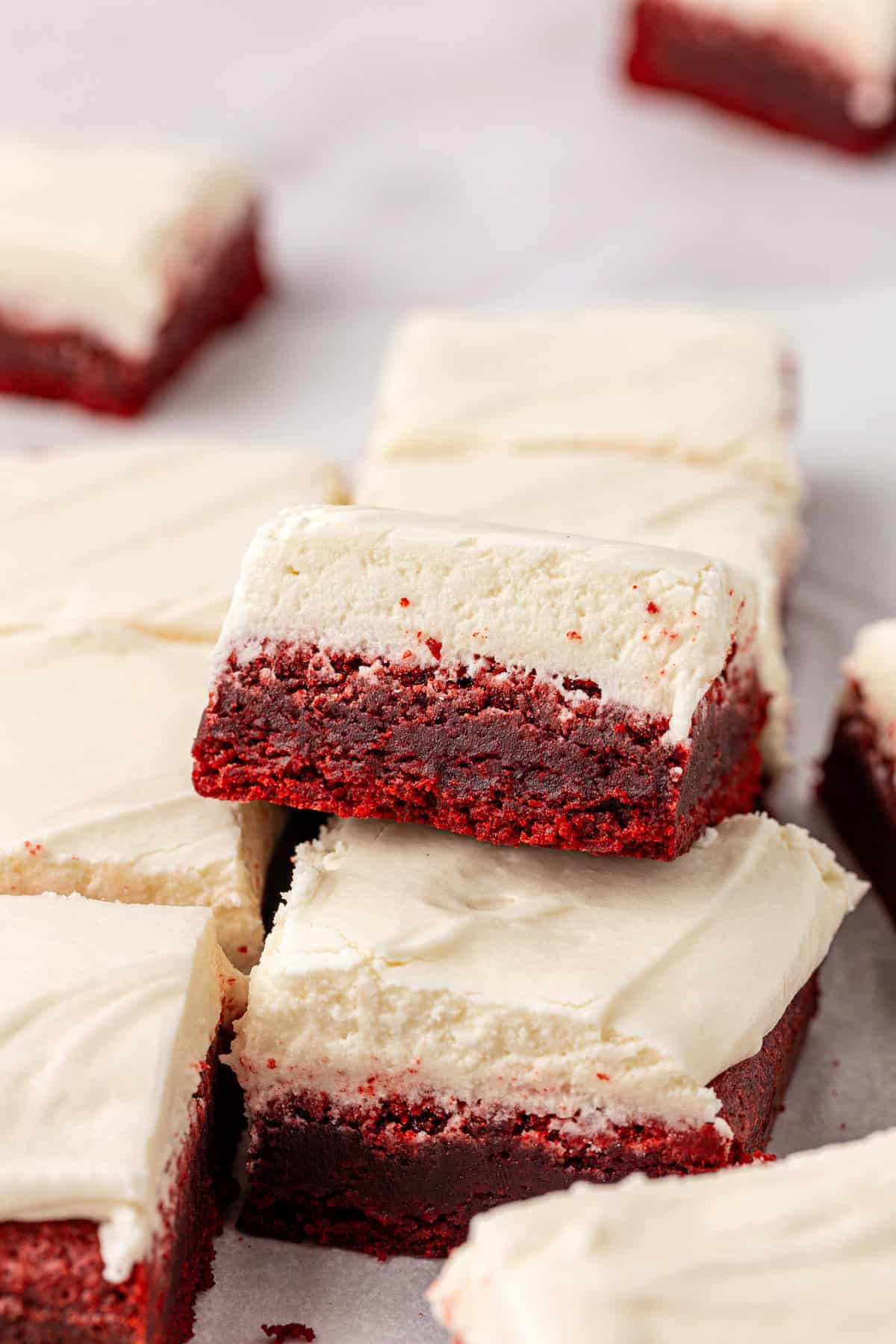 scattered red velvet brownies with one leaning on top of another, showing the two even layers of brownie and cream cheese frosting