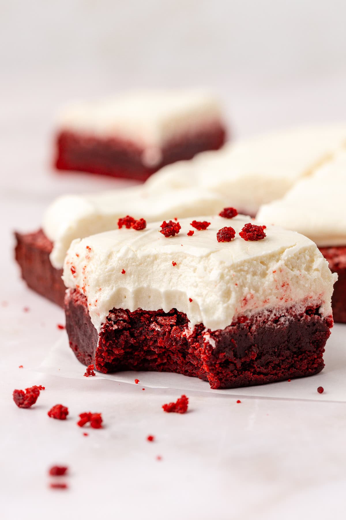 close up of a red velvet brownie topped with cream cheese frosting with a bite missing out of it, sprinkled with red velvet crumbs that are also scattered around