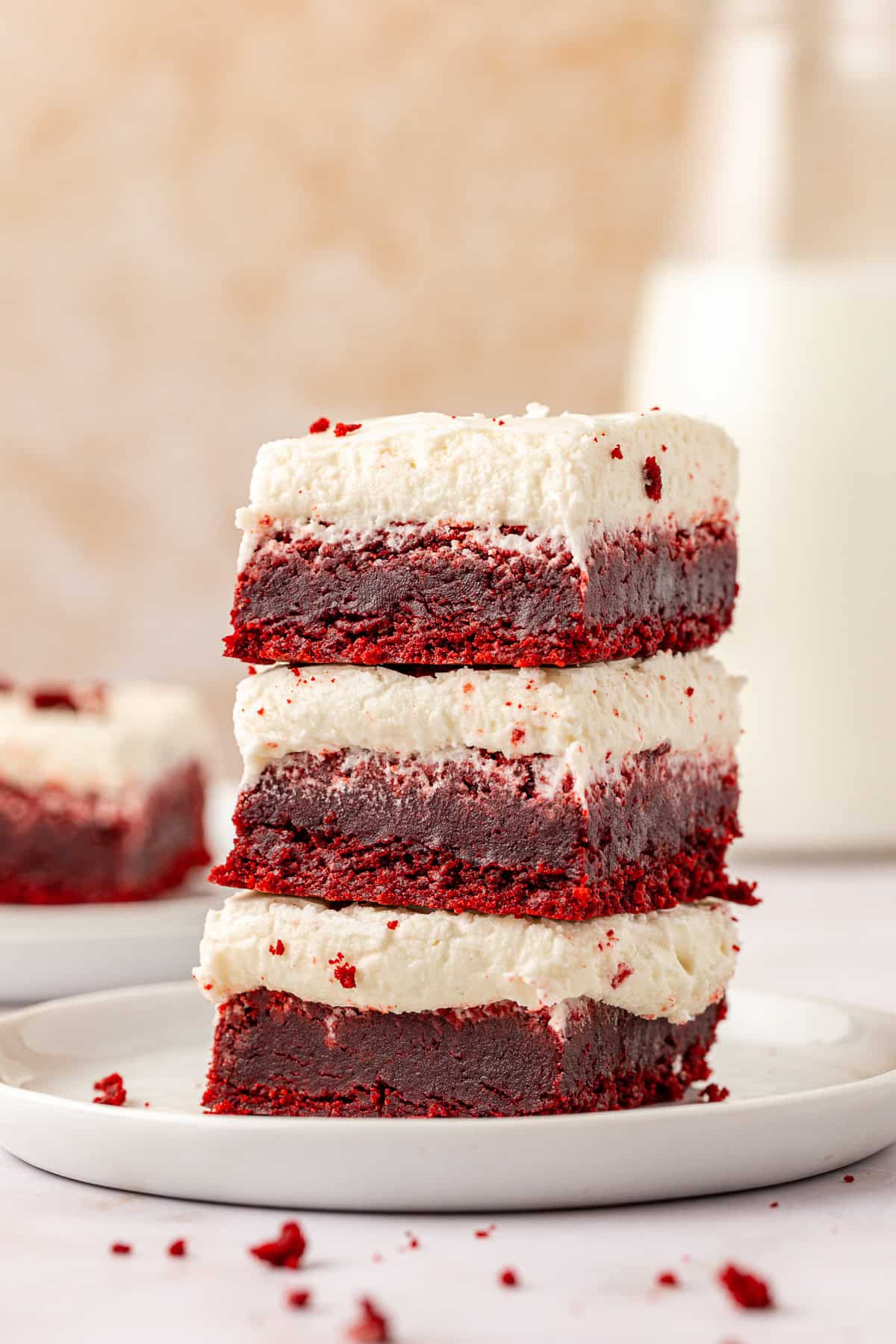 three red velvet brownies topped with cream cheese frosting stacked on a small white plate with red velvet crumbs scattered around