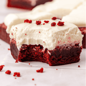 close up of a red velvet brownie topped with cream cheese frosting with a bite missing out of it, sprinkled with red velvet crumbs that are also scattered around