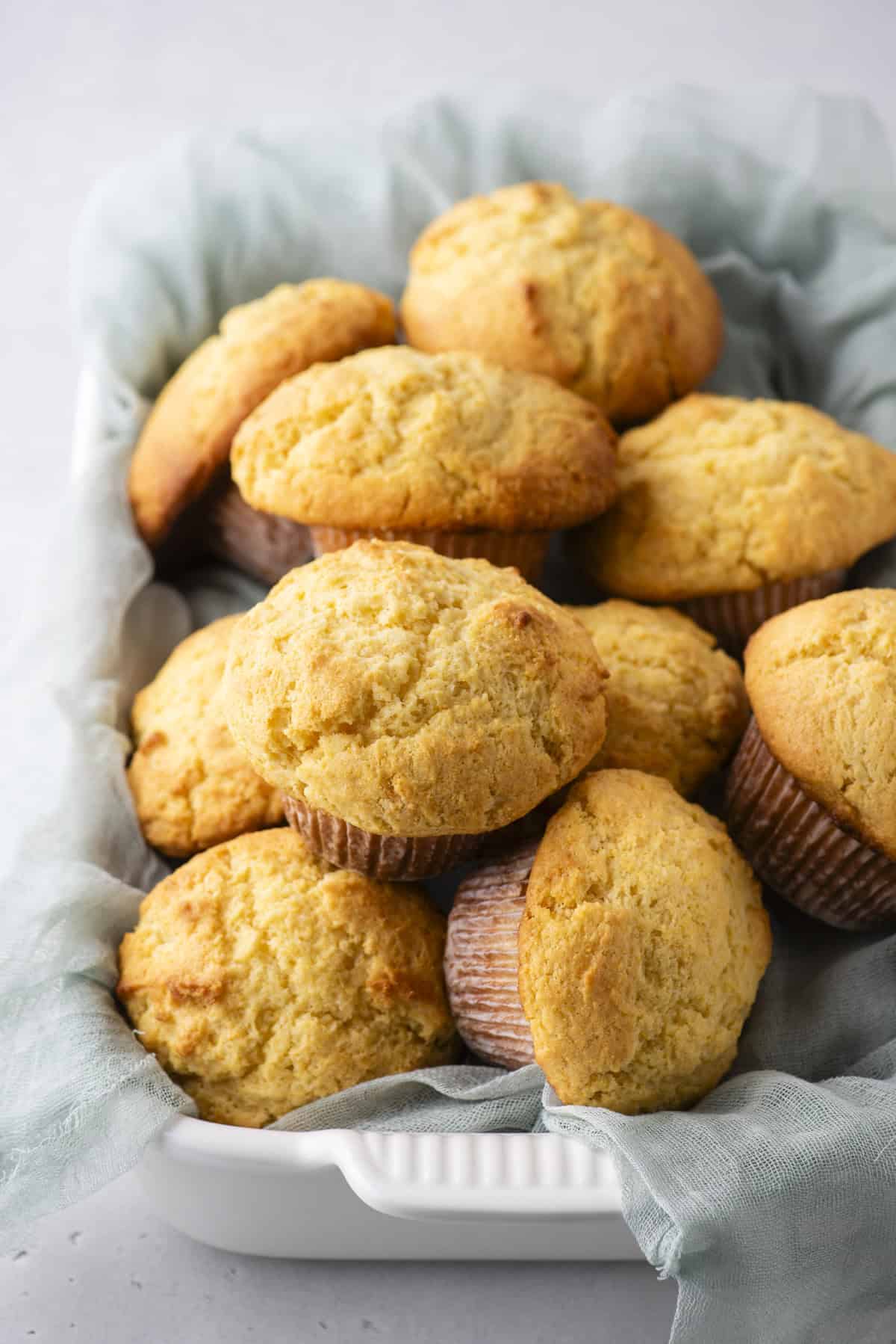 A white rectangular dish holds a stack of golden-brown muffins, lined with a light blue cloth. The muffins are fluffy with slightly cracked tops, suggesting they are freshly baked. The background is a smooth, light gray surface.