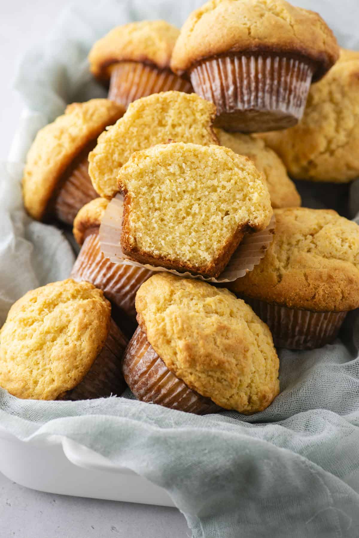 A pile of freshly baked corn muffins is arranged in a white dish lined with a light gray cloth. One muffin is cut in half, revealing its moist, crumbly interior texture.