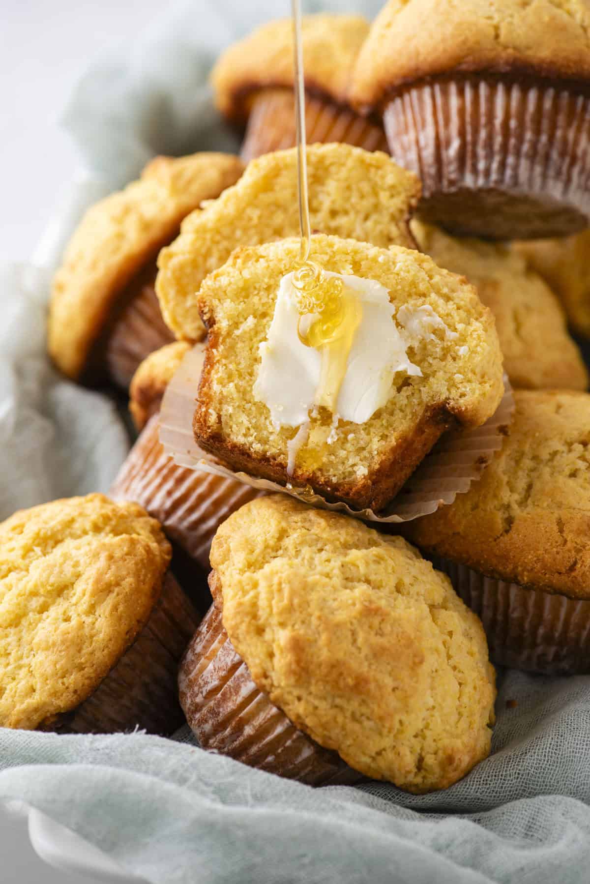 A plate of corn muffins with one muffin cut open, topped with melting butter and drizzled with honey. The muffins are golden-brown with a soft, moist texture. A light blue cloth is underneath the plate.