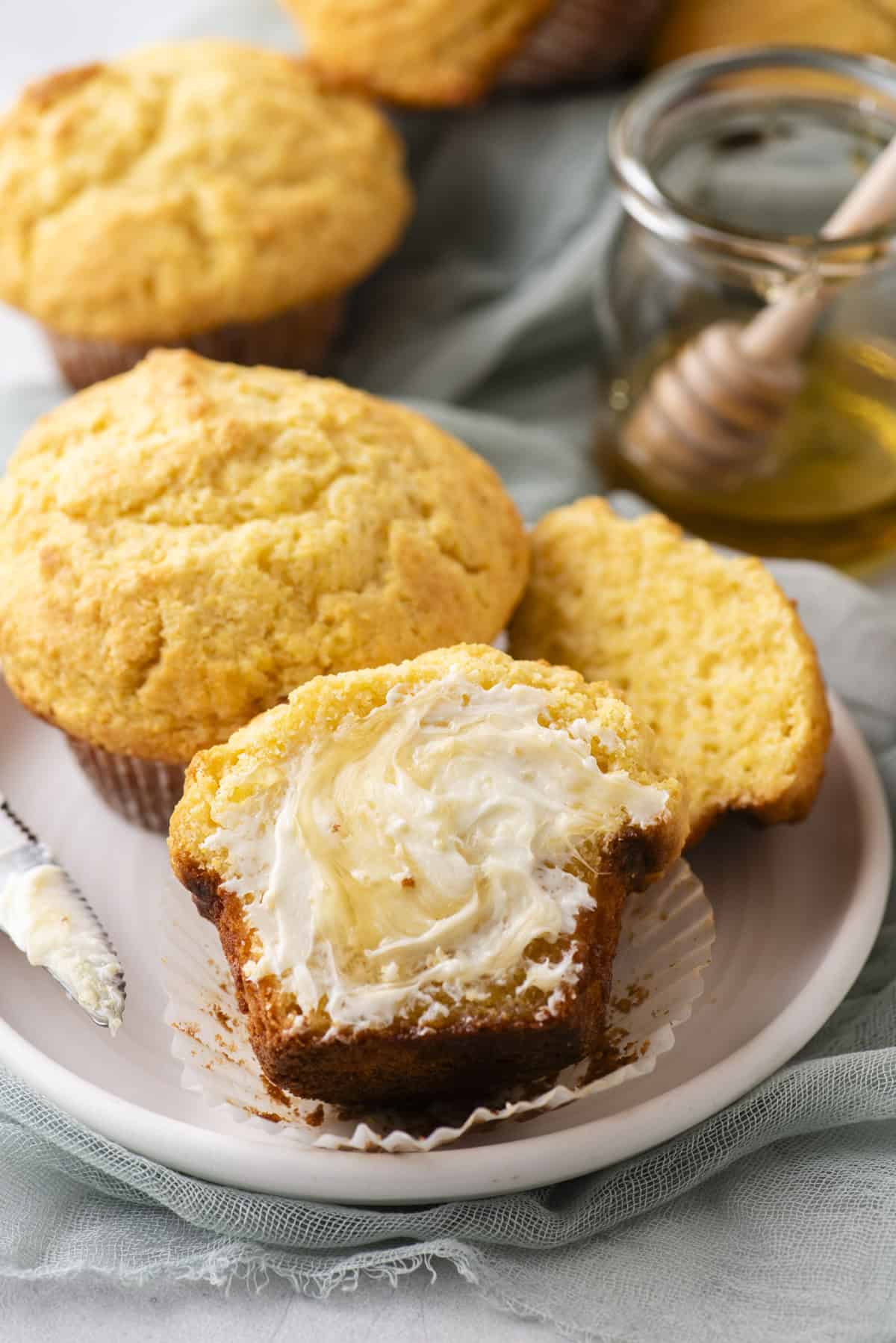 A plate with corn muffins, one cut open and spread with butter. A honey dipper rests on a jar of honey in the background. A green cloth is partially visible under the plate.