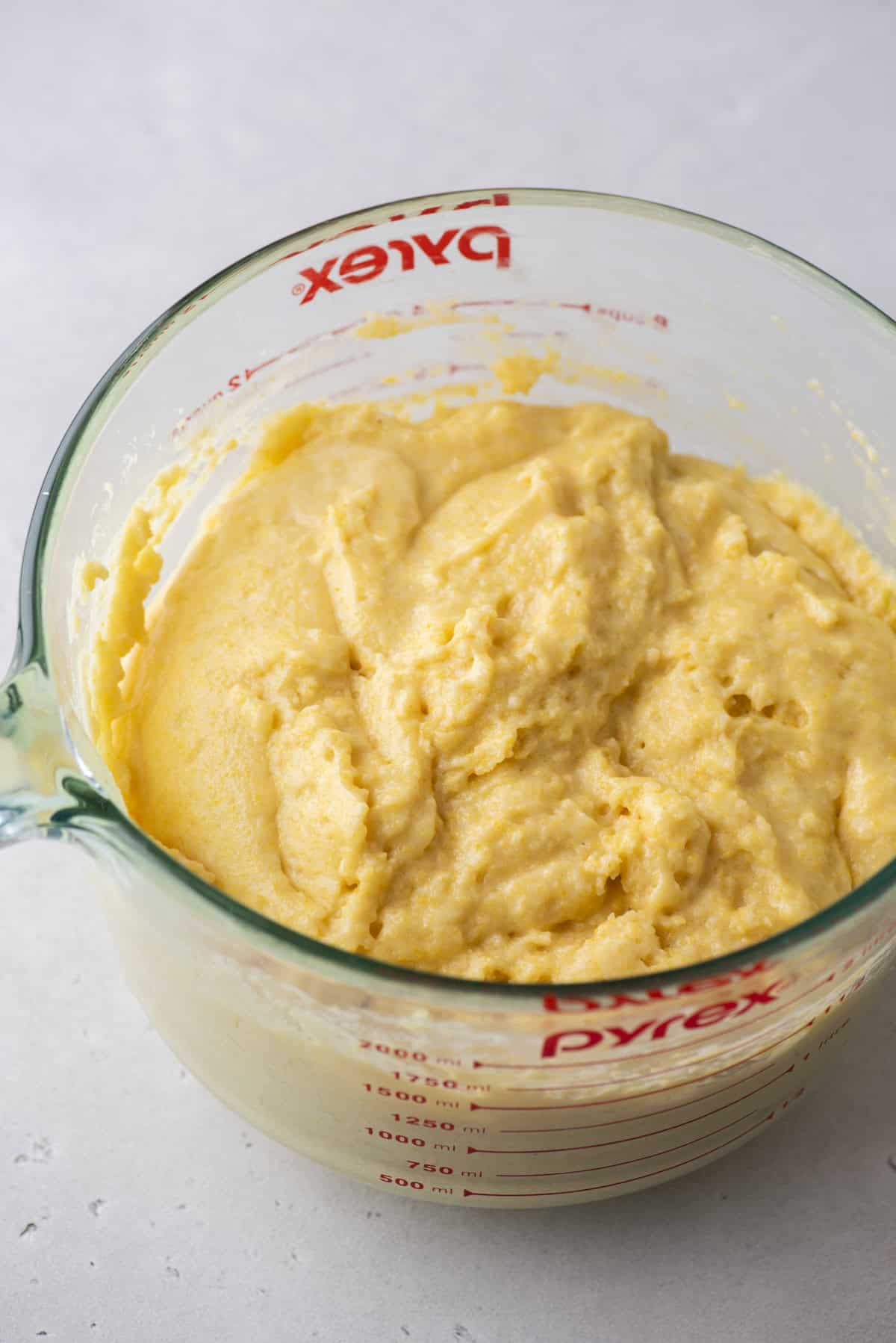 A clear glass measuring cup filled with light yellow, thick cornbread batter. The sides of the cup display red measurement markings. The background is a light-colored surface.