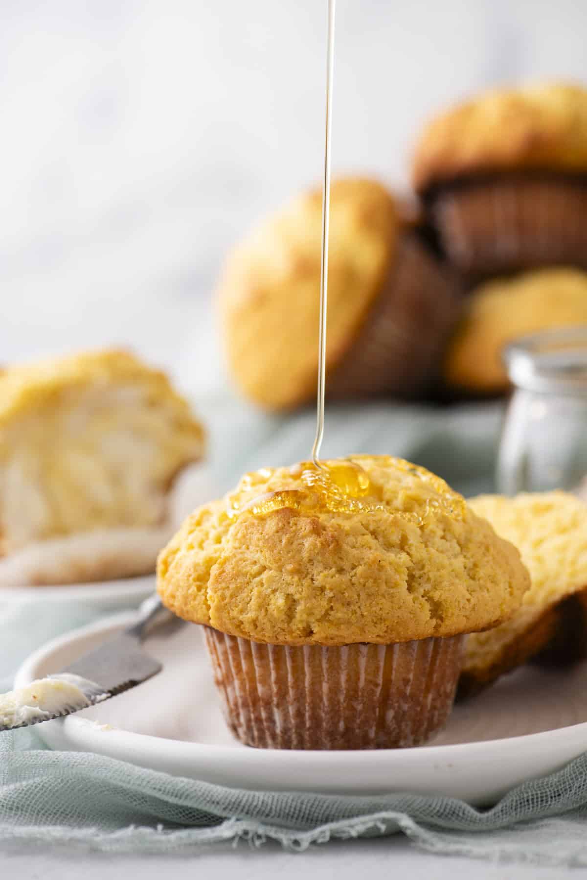 A muffin is on a white plate, being drizzled with honey. The background shows more muffins and a buttered muffin half. A small jar of honey is nearby. The scene is set on a light blue cloth and a marble surface.