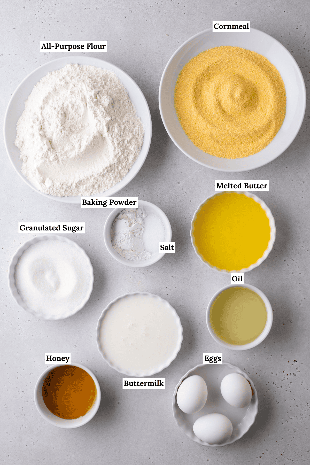 A collection of corn muffin ingredients in bowls on a countertop: all-purpose flour, cornmeal, granulated sugar, salt, baking powder, melted butter, oil, honey, buttermilk, and three eggs. Each item is labeled with text.