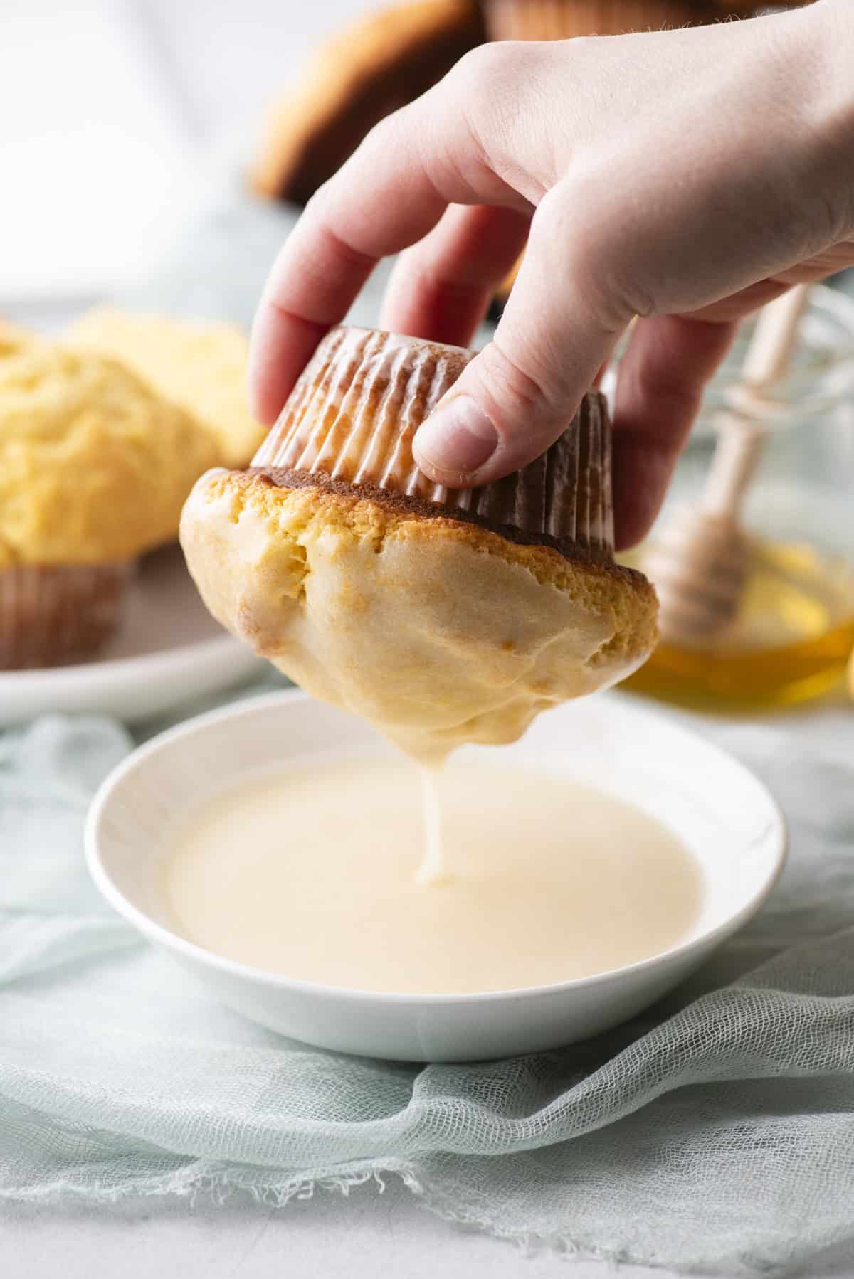 A hand dipping a muffin with a corrugated wrapper into a bowl of creamy glaze. More muffins are visible on a plate in the background, alongside a jar with a honey dipper. A soft, light blue cloth lies beneath.