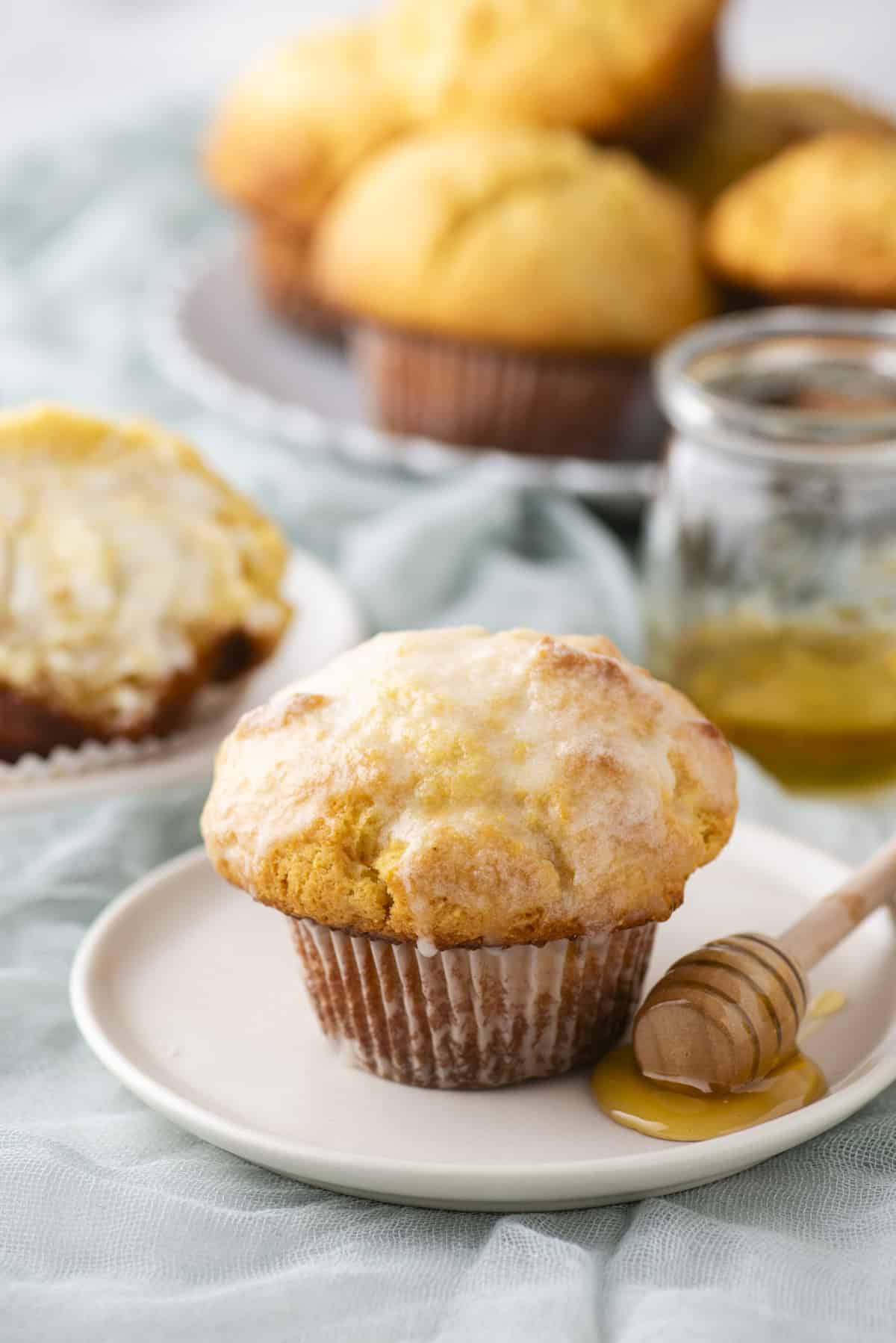 A muffin sits on a white plate with a honey dipper nearby. The muffin is golden brown with a light glaze. In the background, theres a partially eaten muffin on another plate and more muffins on a tray, with a jar of honey beside them.