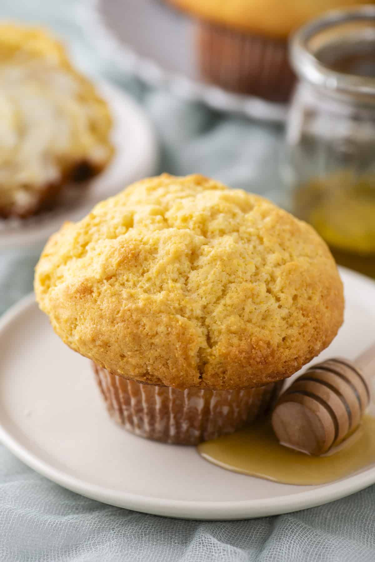 A golden-brown muffin on a white plate with a wooden honey dipper coated in honey beside it. The background shows a glass jar of honey and another plate with a partially visible muffin. The setting conveys a cozy, homemade feel.