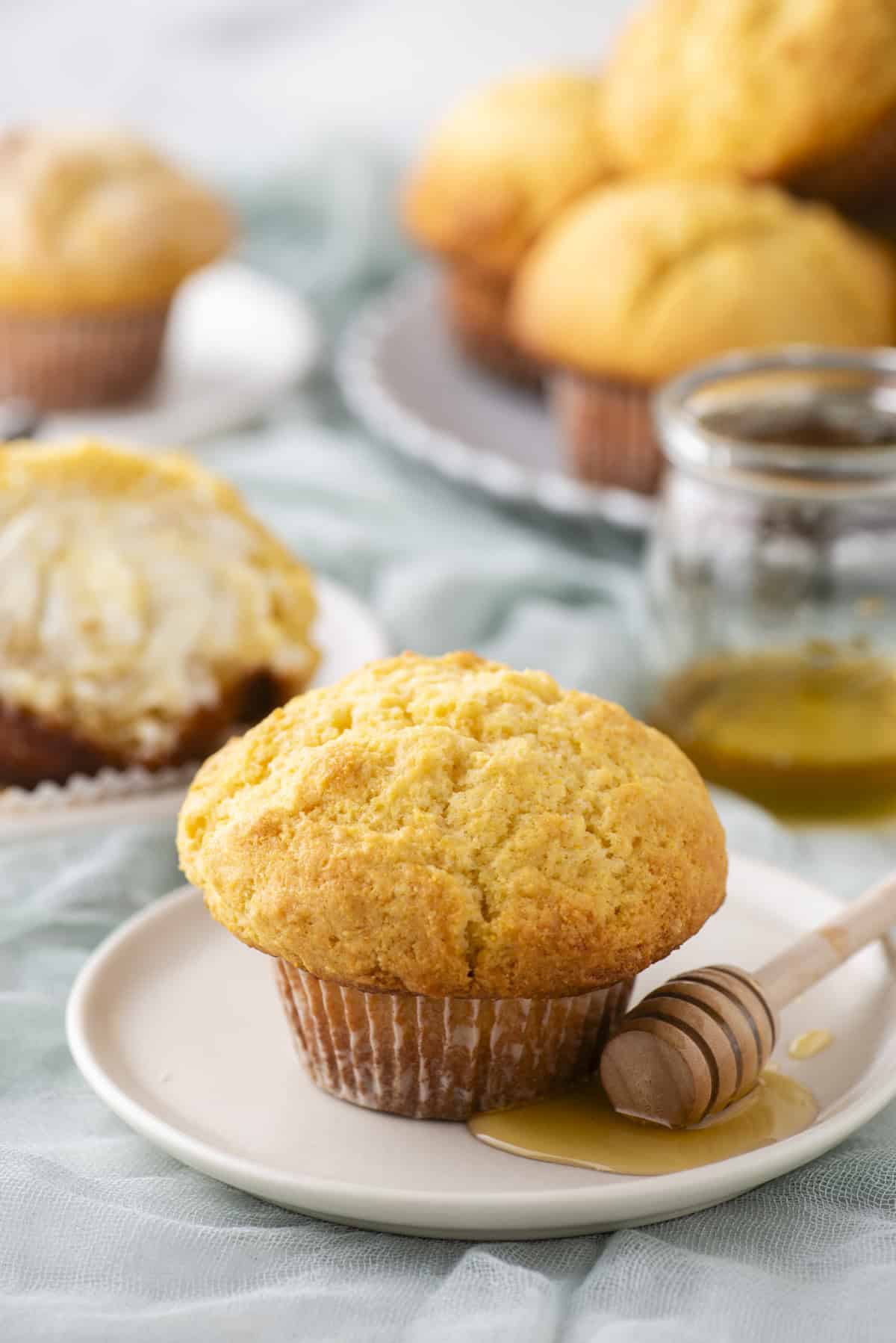 A cornbread muffin sits on a plate with a wooden honey dipper and honey drizzled nearby. In the background, more muffins are stacked on a plate, and one is cut open with butter. A jar of honey is also visible.