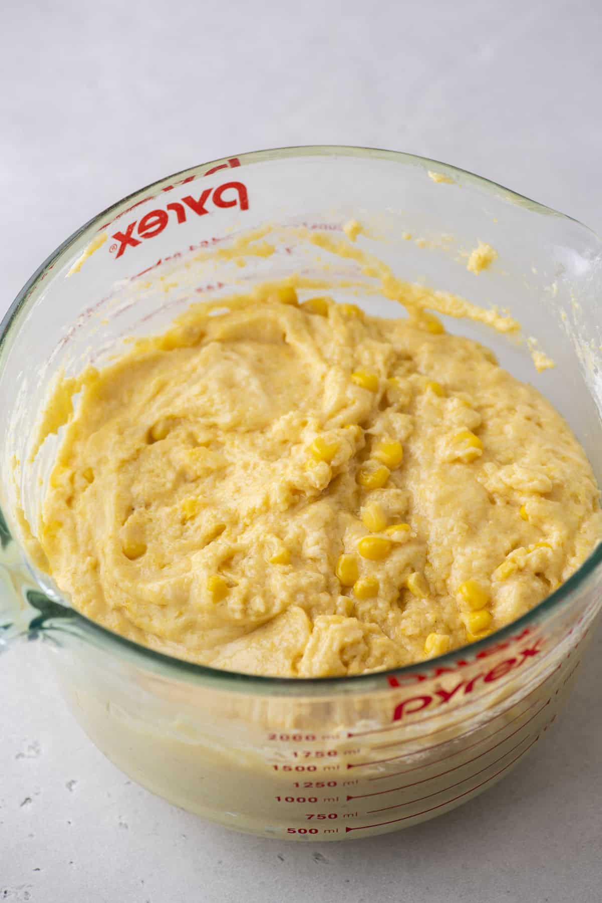 A glass Pyrex measuring cup brims with creamy yellow corn muffin batter, speckled with pieces of corn. Resting on a light-colored surface, the cup’s red measurement markings stand out against the glass.