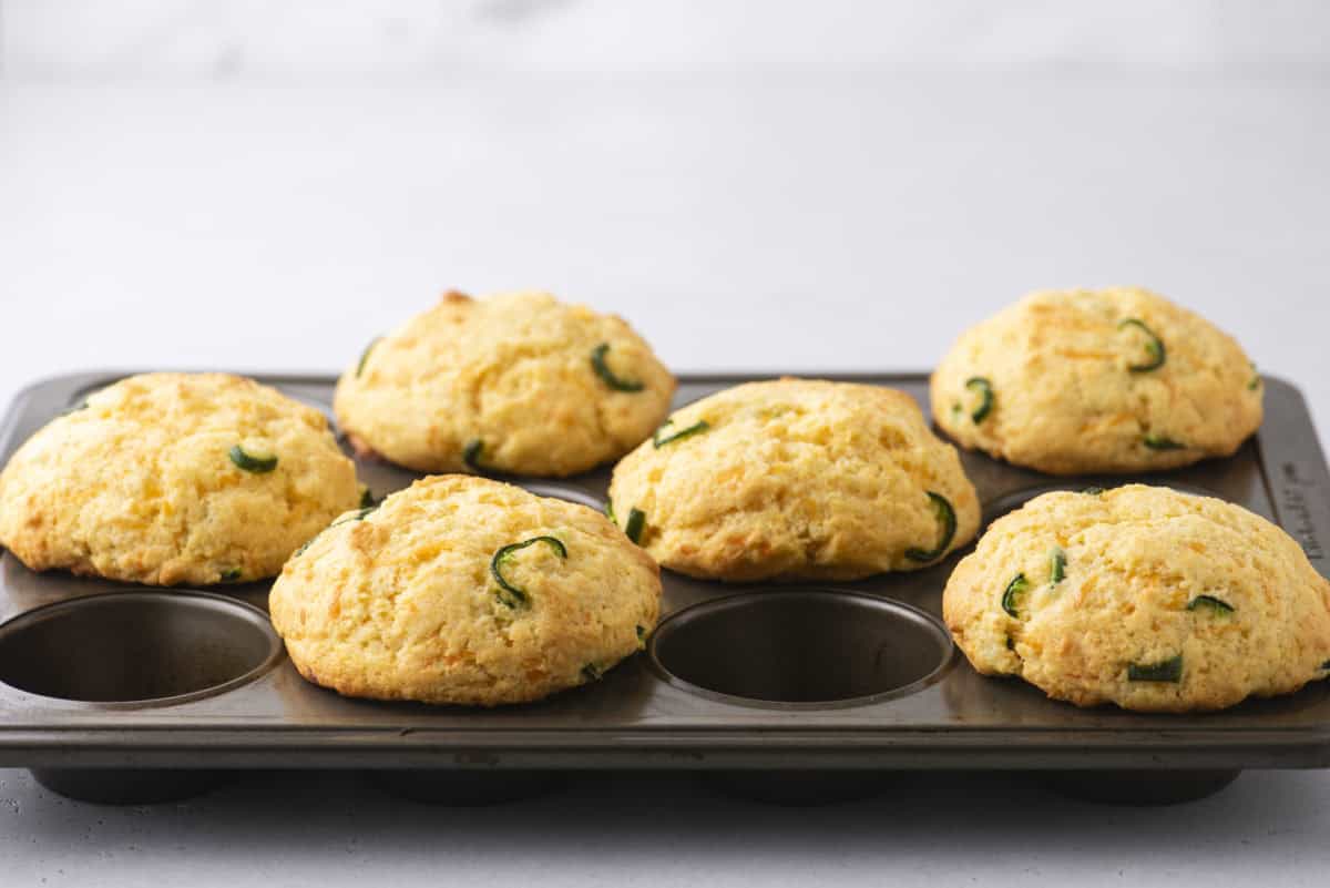 A muffin tin showcasing six freshly baked corn muffins, each adorned with slices of green jalapeños, sits invitingly. The golden brown corn muffins contrast beautifully against the light gray background.