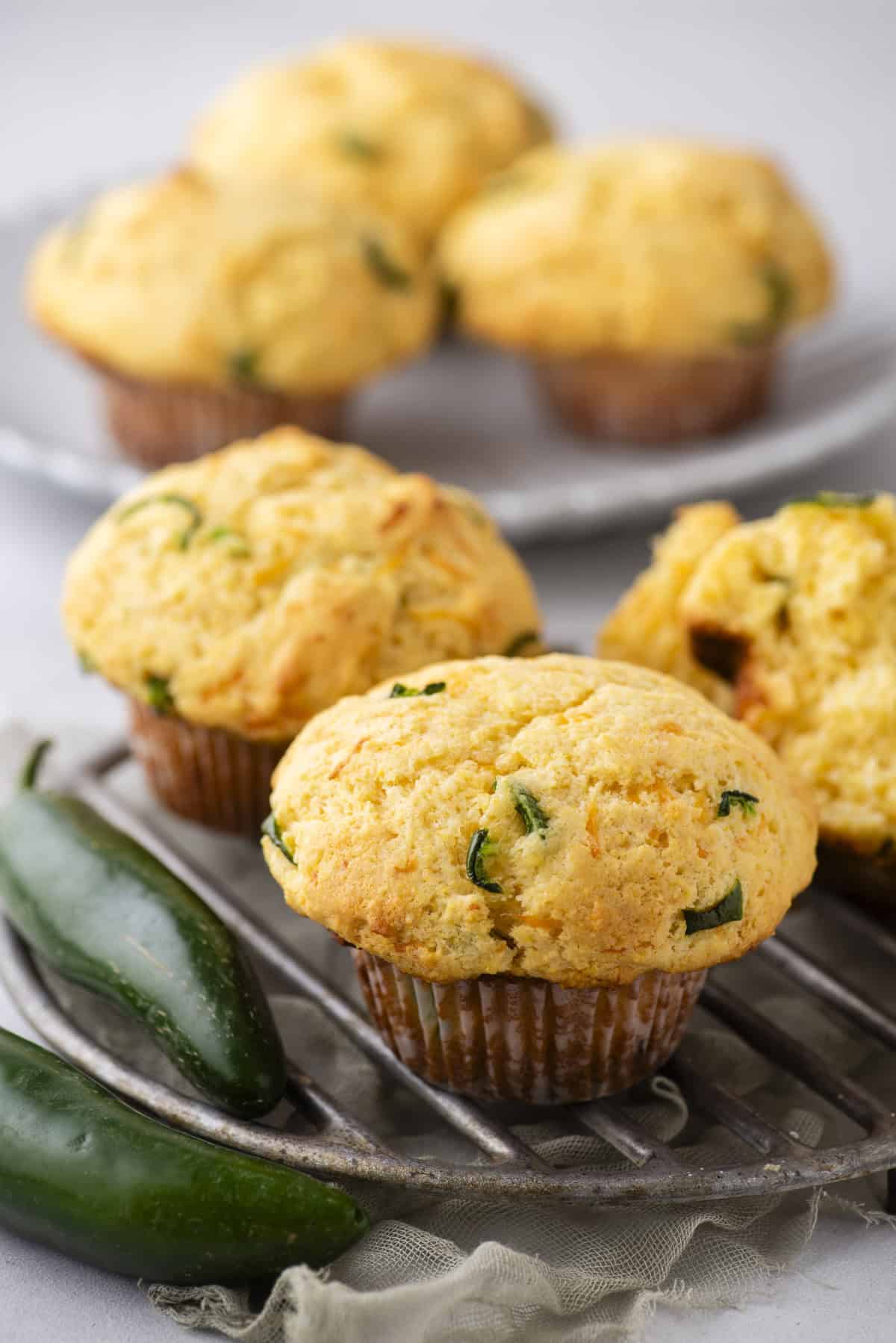 Close-up of freshly baked corn muffins with jalapeño slices on a cooling rack. Two green jalapeños rest nearby. A blurred plate with more muffins is visible in the background.