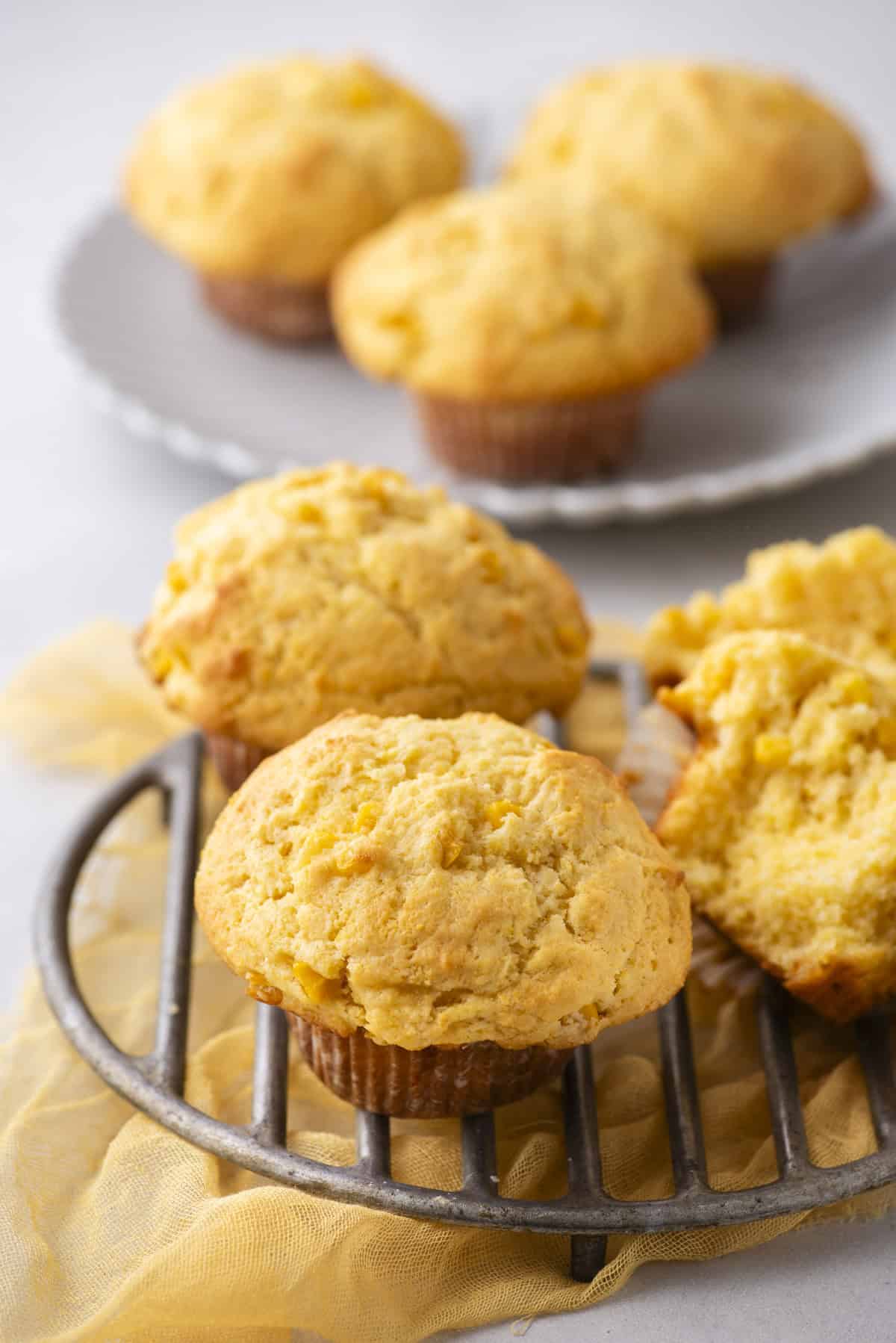 Freshly baked corn muffins rest on a cooling rack, with one split open to reveal its moist texture. A plate adorned with more corn muffins sits in the background, all set upon a light-colored cloth that adds to the inviting ambiance.