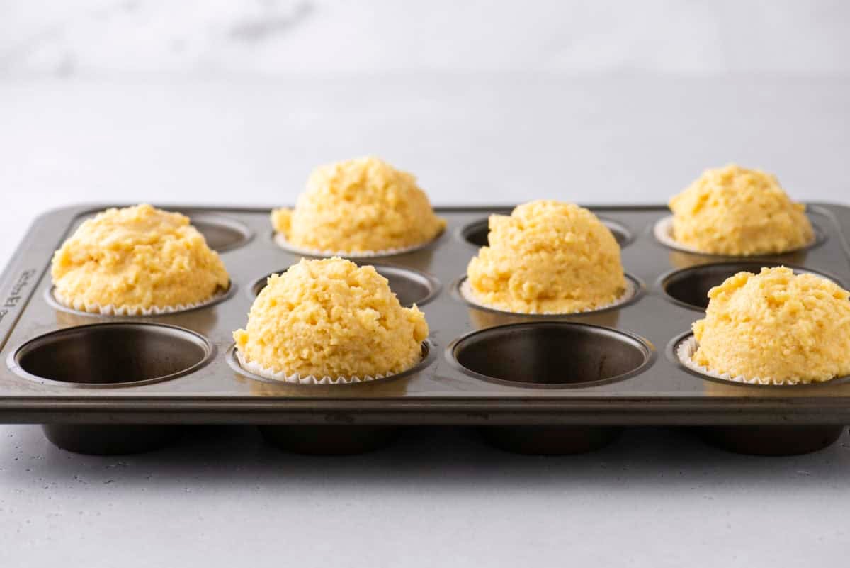 A muffin tray with seven scoops of uncooked corn muffin batter in paper liners, placed on a light gray countertop. The dough is rough and slightly mounded above the tray. The background is softly blurred.