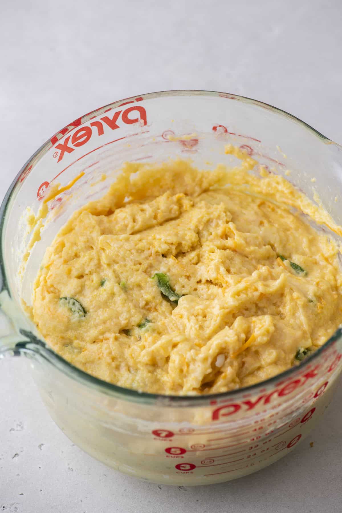 A clear measuring cup brimming with cornbread batter reveals bits of jalapeños mixed in, ready to be transformed into spicy corn muffins. The cup rests on a pristine white countertop.