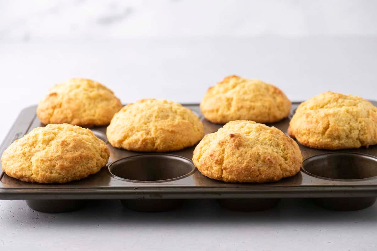 A muffin tray holds six golden-brown muffins, placed on a light-colored countertop. The muffins are slightly domed and appear freshly baked, with a soft, textured surface.