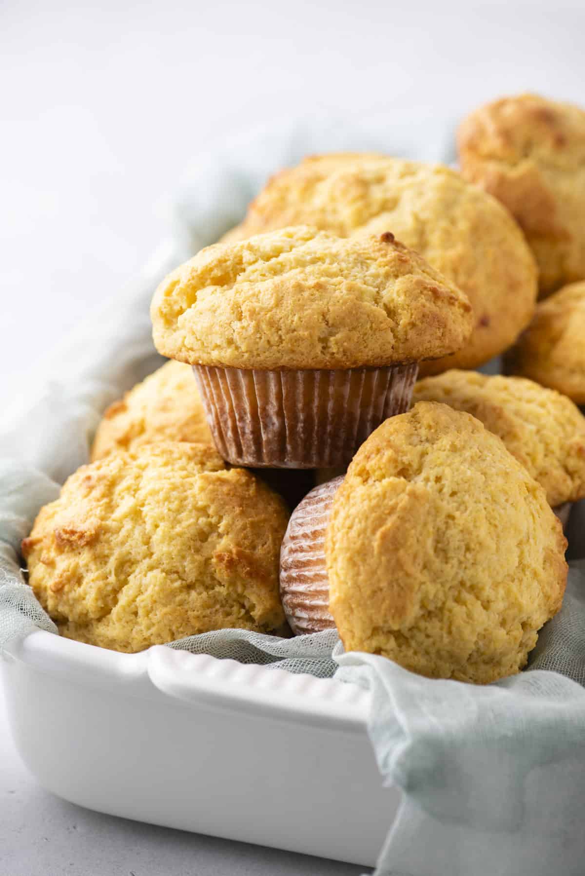 A tray filled with golden-brown muffins, sitting on a light blue cloth. One muffin is prominently placed on top, showing its textured surface and wrapped in a paper liner. The background is softly blurred.