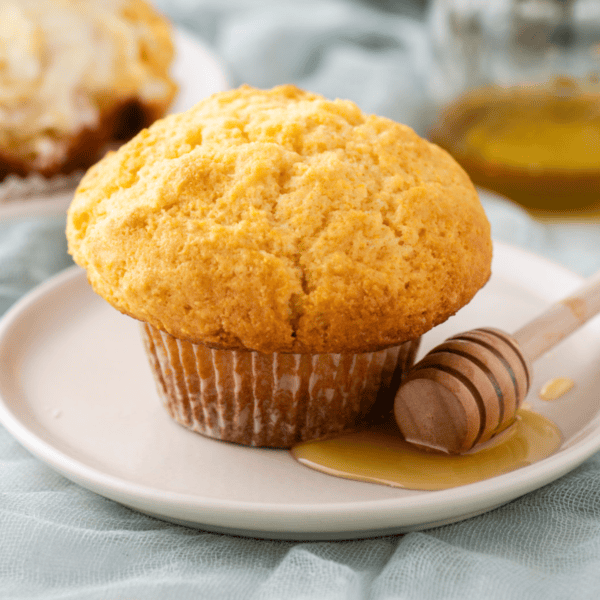 A golden-brown muffin sits on a white plate, accompanied by a honey dipper with honey pooling beside it. The background features a soft, light blue fabric and a blurred second plate and glass jar.