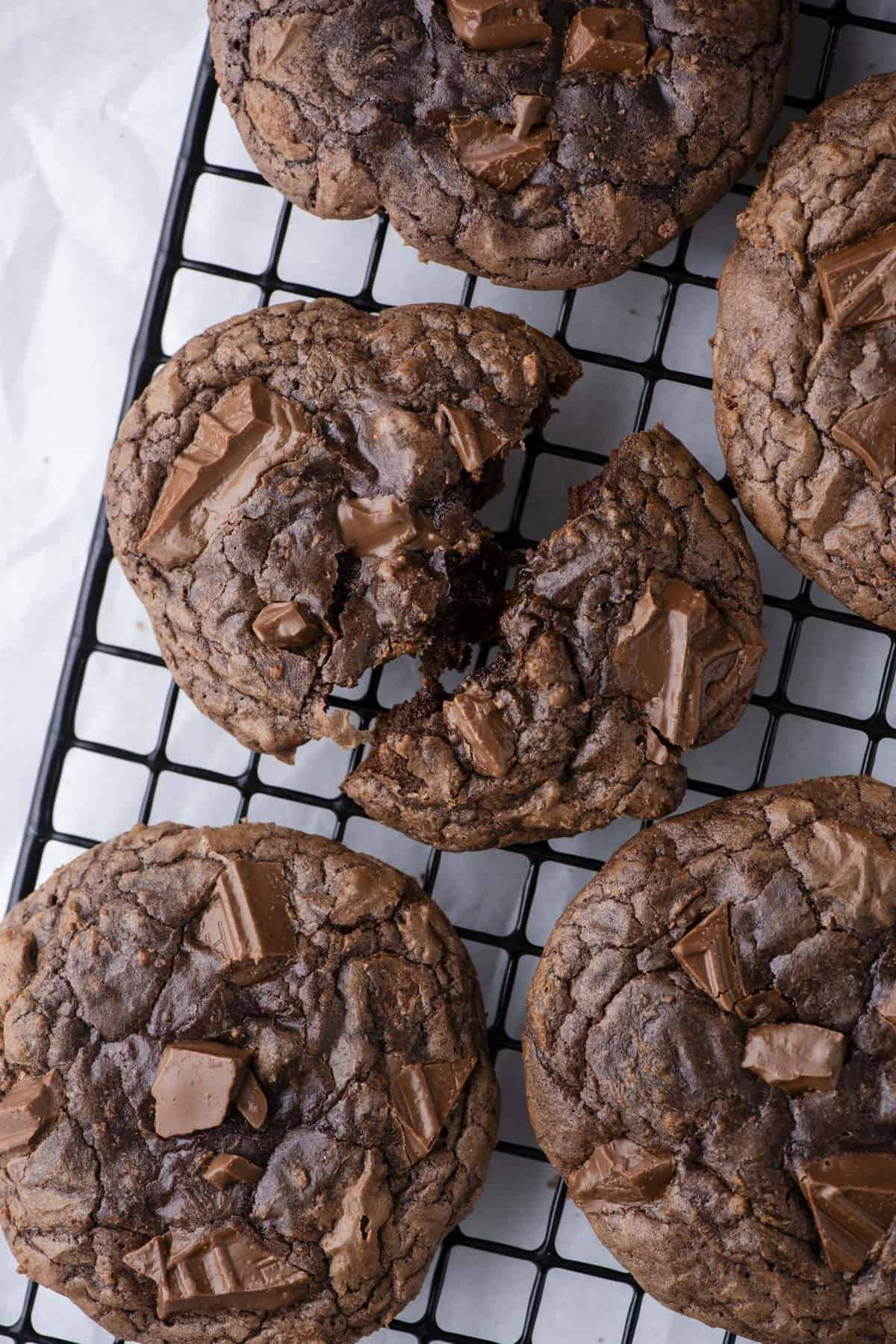 Four large, chunky chocolate cookies with chocolate chunks rest on a black wire cooling rack. One cookie is broken in half, revealing a soft, gooey inside. The cookies are on a white parchment paper background.