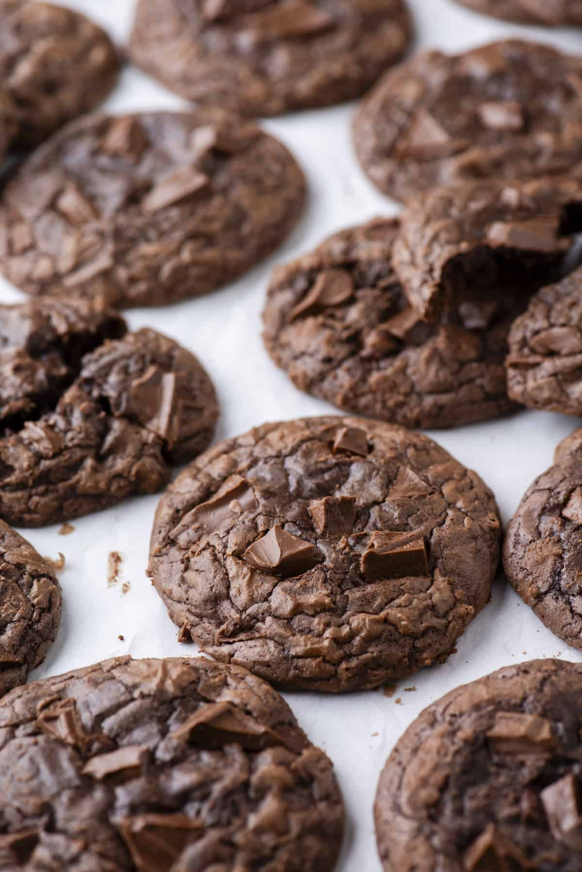 A close-up of several rich, chocolate cookies with chunks of chocolate on top, arranged on white parchment paper. Some cookies are broken, revealing their soft, fudgy texture inside.