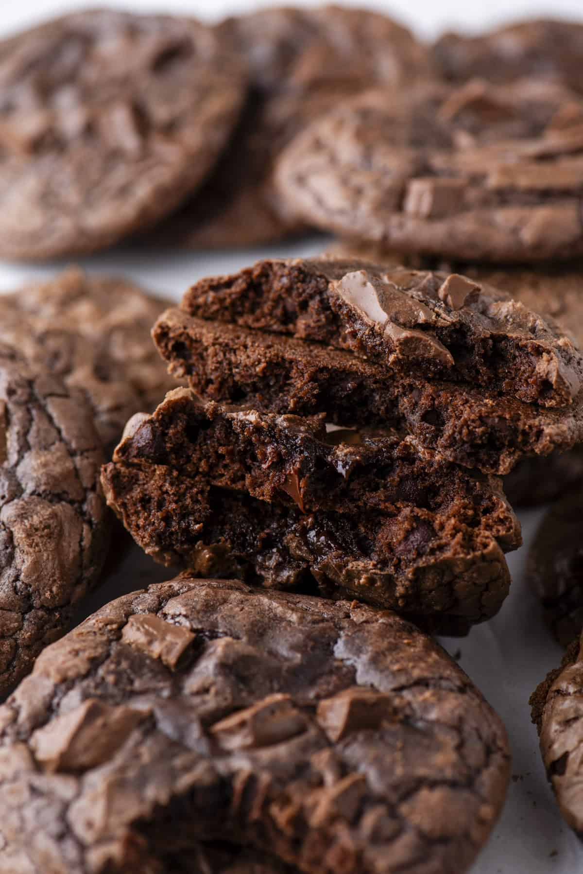 A close-up of rich, chocolate cookies with chunks of chocolate, including one cookie broken in half to reveal a gooey, fudgy center. Other whole cookies are visible in the background.
