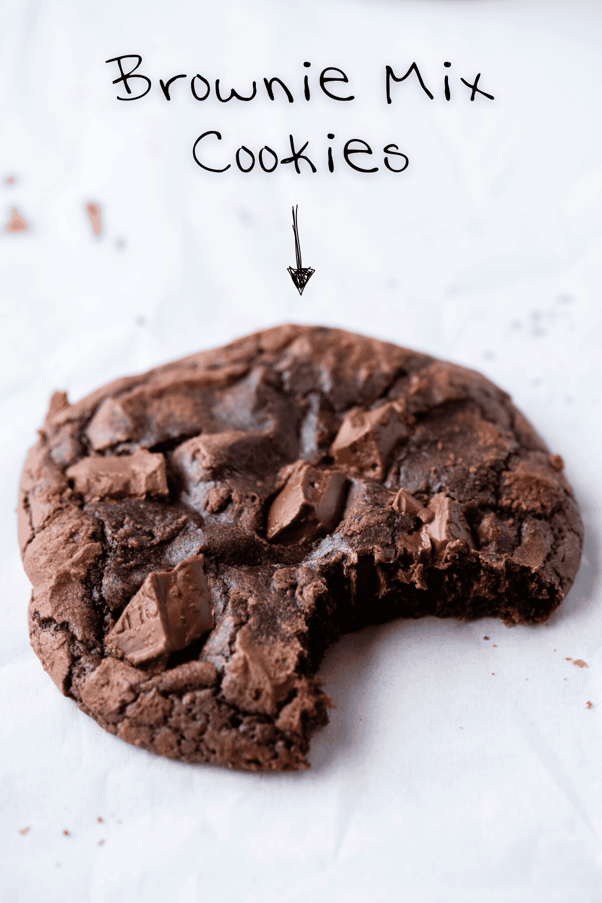 A chocolate brownie cookie with chunks of chocolate and a bite taken out, sits on white parchment paper. Above it, handwritten text reads “Brownie Mix Cookies” with an arrow pointing to the cookie.