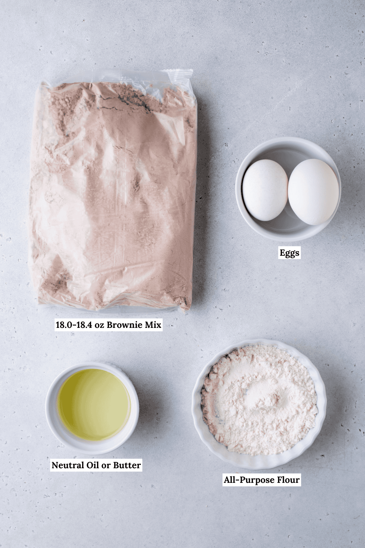Top-down view of four labeled ingredients for brownies: a bag of brownie mix, two eggs in a bowl, a small bowl of neutral oil or butter, and a small bowl of all-purpose flour on a light surface.