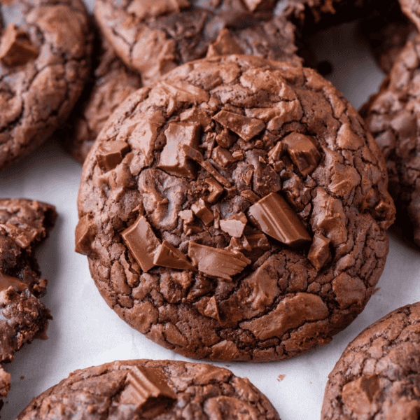 Close-up of several rich, fudgy chocolate cookies topped with chunks of chocolate, displayed on a white surface.