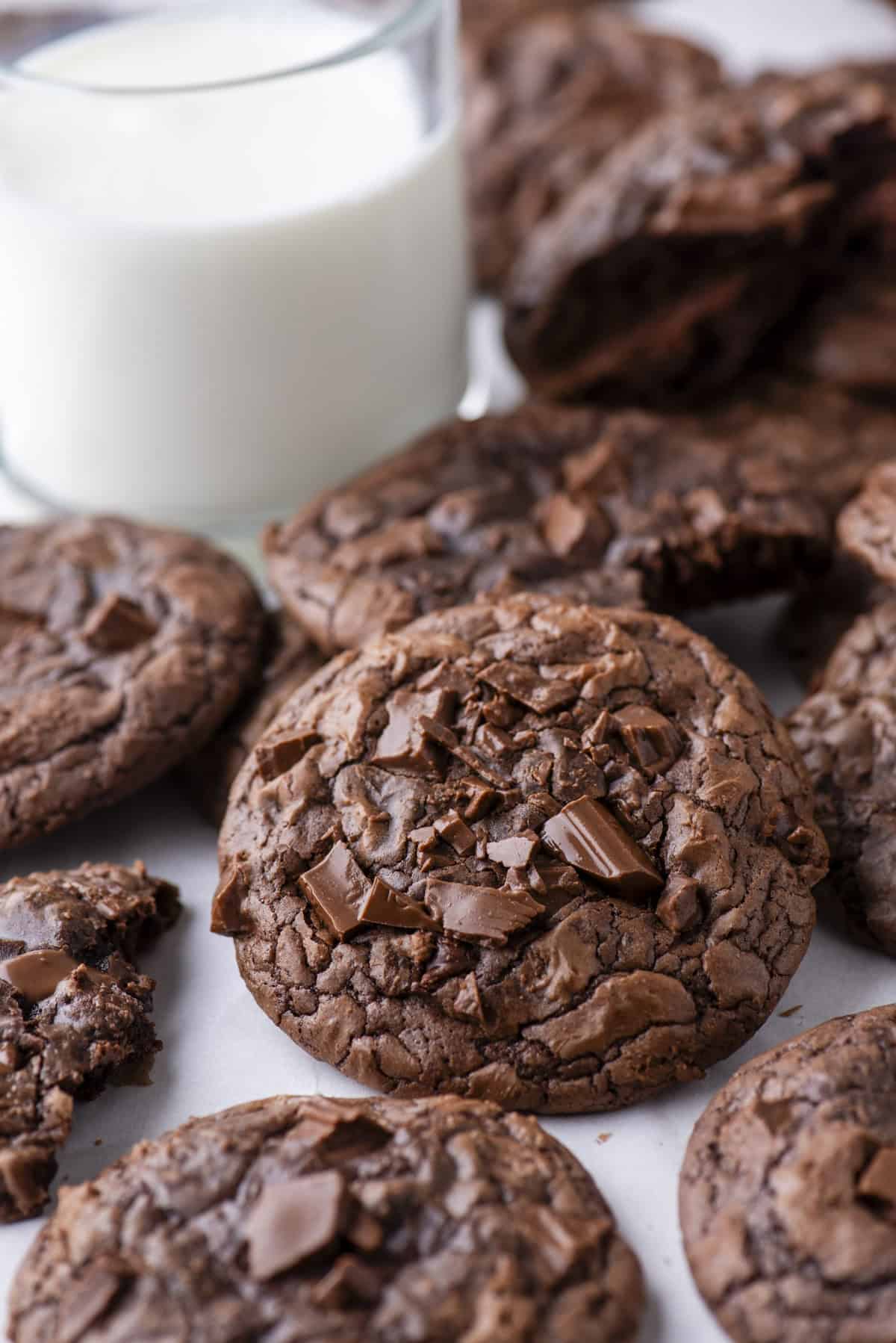 A close-up of several chocolate chip cookies with chunks of chocolate on top, arranged on a white surface next to a glass of milk.
