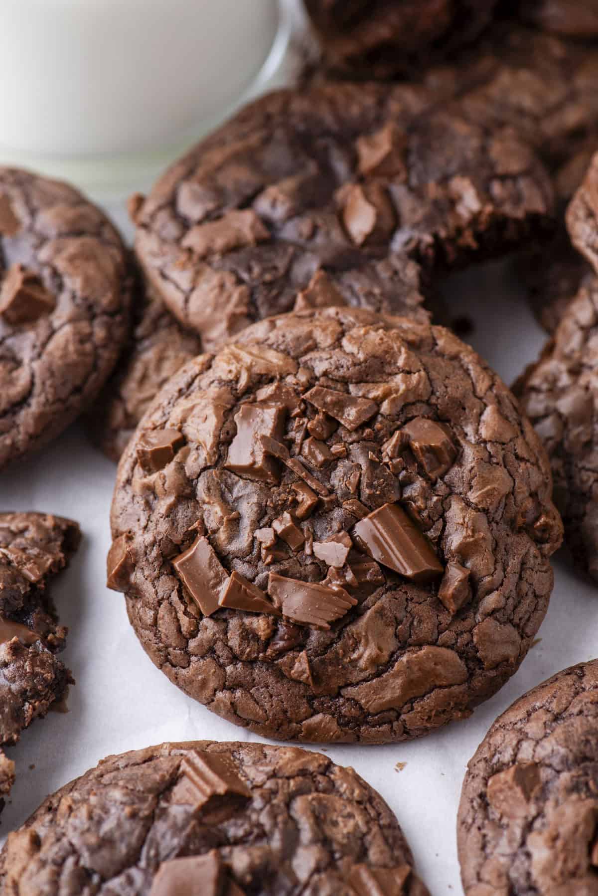 A close-up of several rich, fudgy chocolate cookies with chunks of chocolate on top, arranged on a white surface. A glass of milk is partially visible in the background.