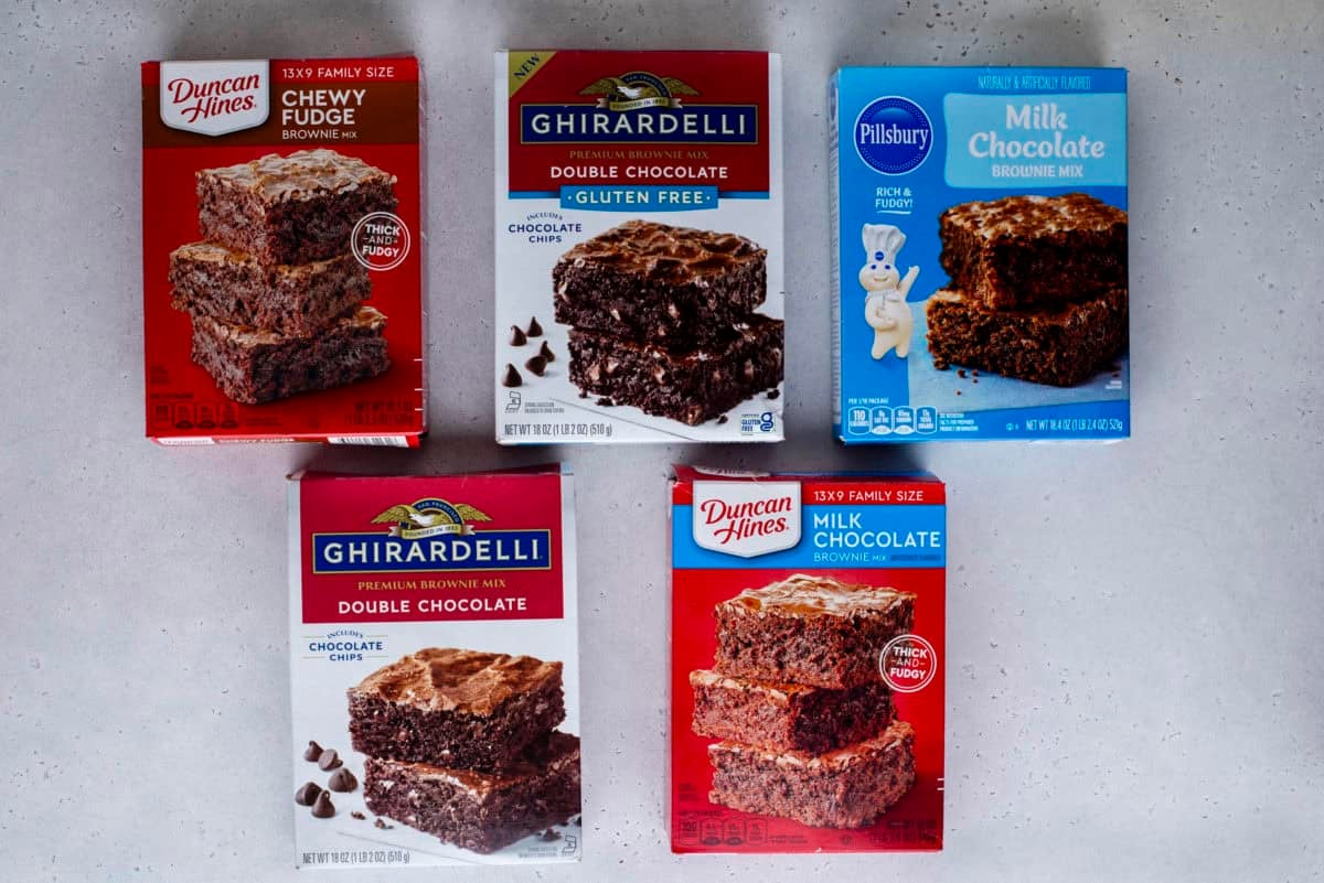 Five boxes of brownie mix from Duncan Hines, Ghirardelli, and Pillsbury are arranged on a light gray surface, featuring flavors like chewy fudge, double chocolate, and milk chocolate.