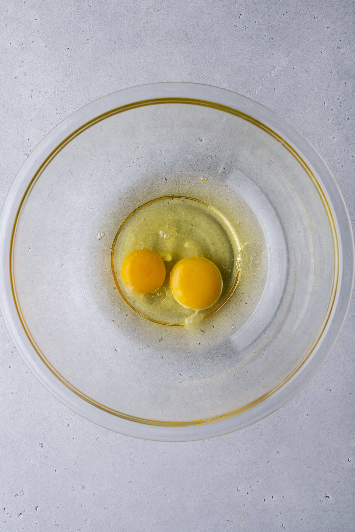 Two raw eggs with unbroken yolks in a clear glass bowl, placed on a light gray textured surface, viewed from above.