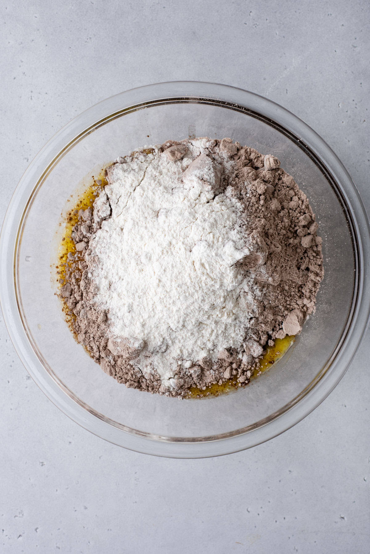 A glass mixing bowl containing unmixed brownie ingredients: cocoa powder, flour, and melted butter on a light gray countertop.