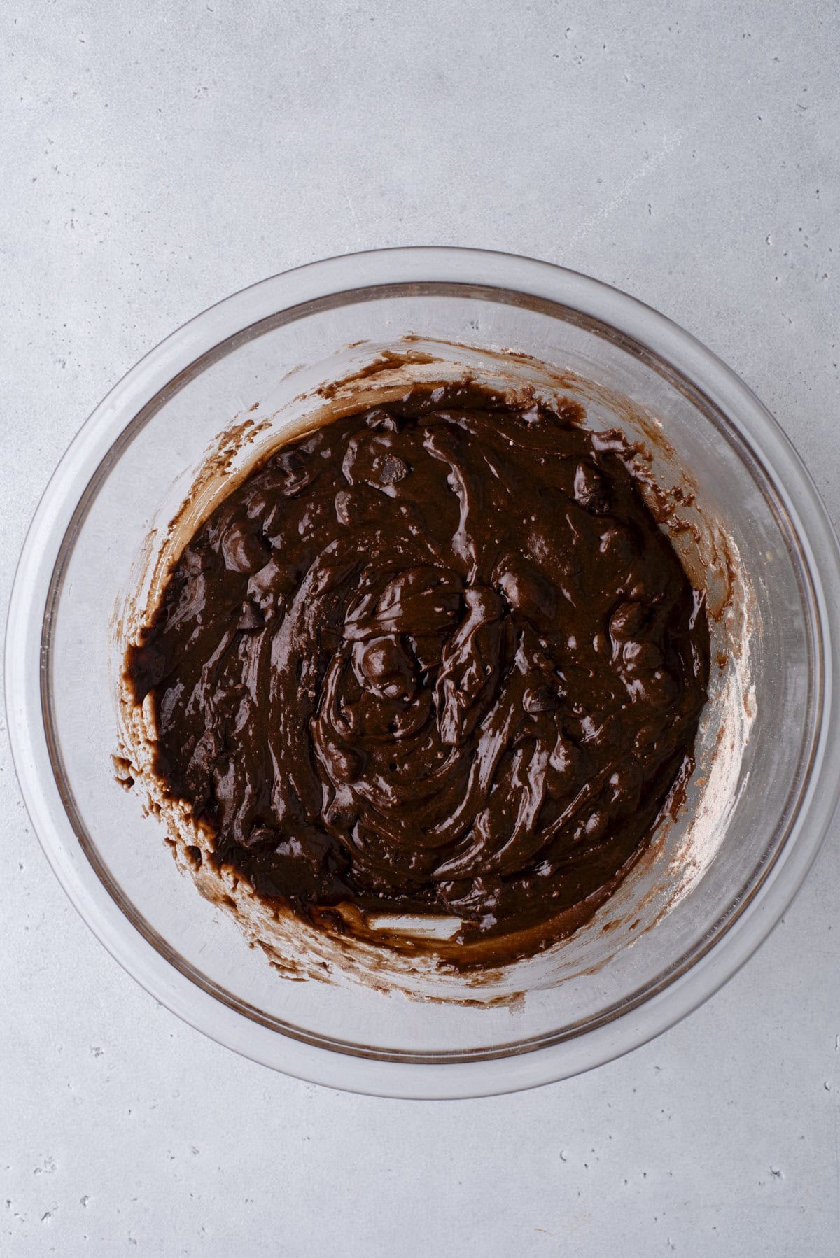 A glass mixing bowl filled with thick, dark chocolate batter sits on a light gray surface. The batter appears to be ready for baking, with some streaks visible on the sides of the bowl.