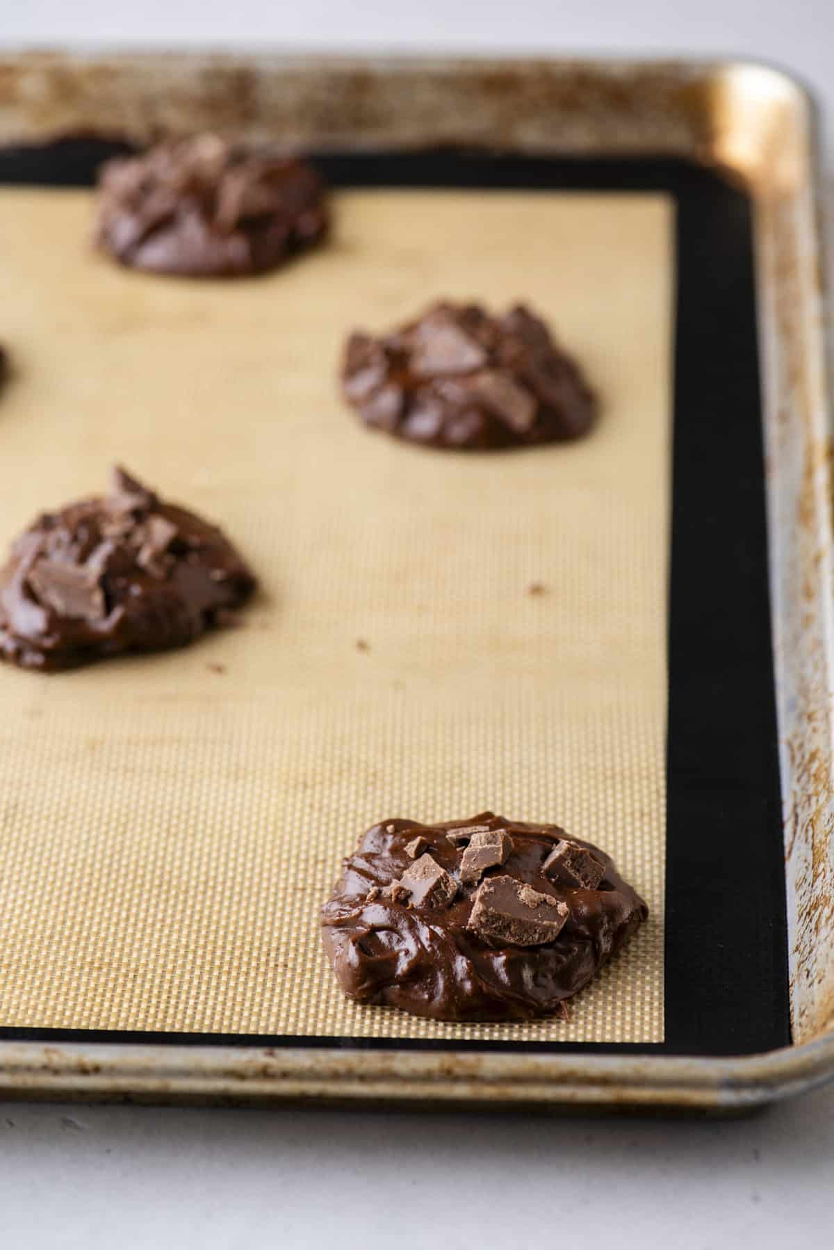 Scoops of chocolate cookie dough with chocolate chunks on a silicone baking mat, spaced apart on a metal baking sheet, ready to be baked.