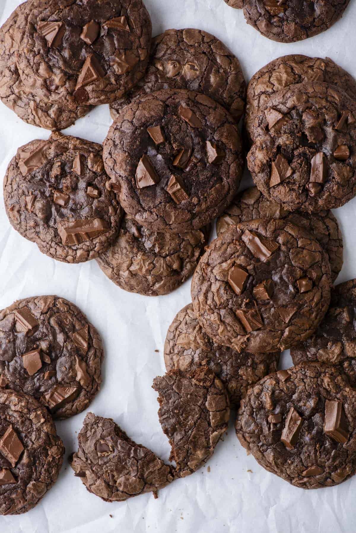 A batch of chocolate cookies with chocolate chunks is arranged on white parchment paper. One cookie is broken in half, revealing a soft, rich interior. The cookies have a crinkled surface and glossy chocolate pieces.