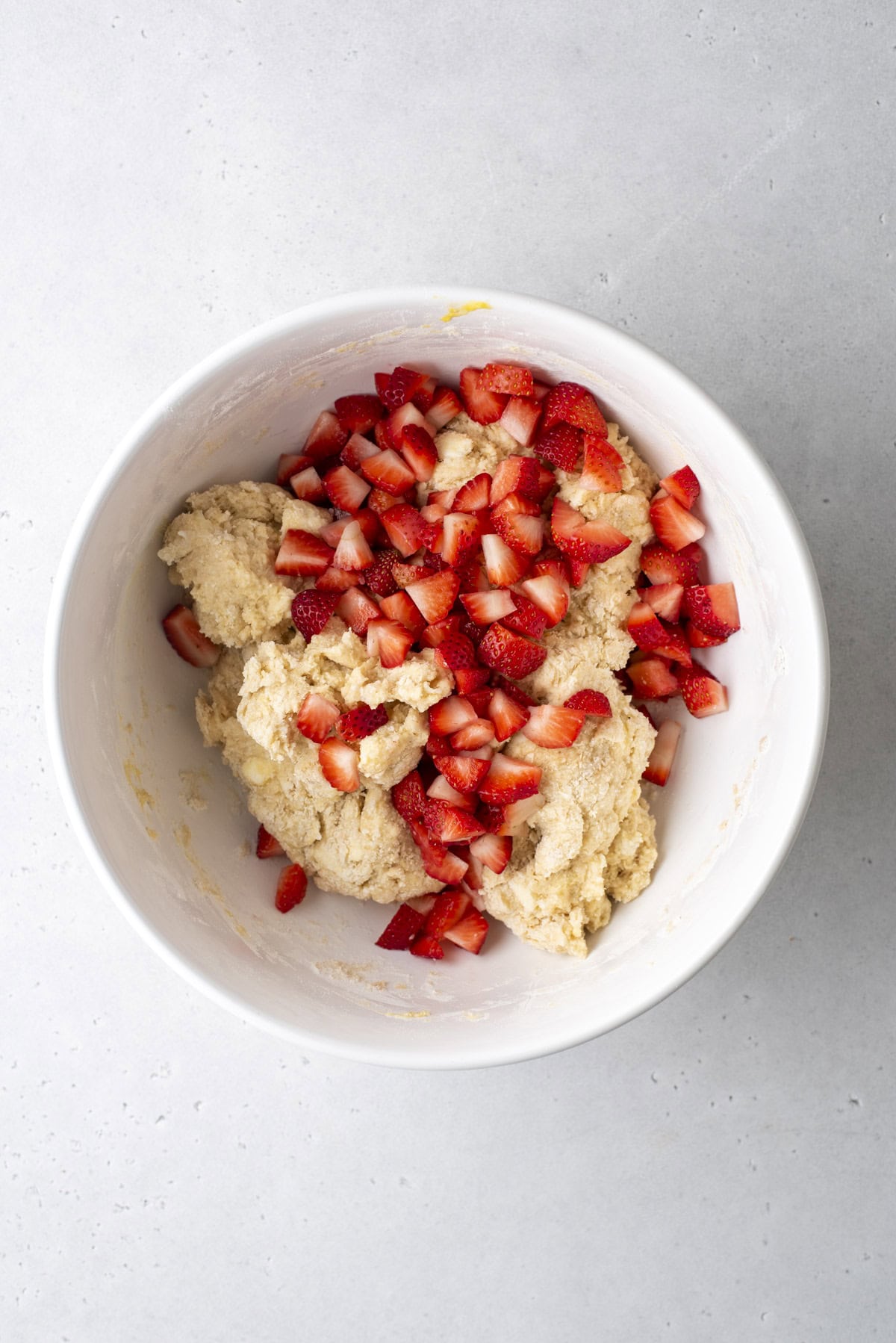 A white bowl contains dough and chopped strawberries on a light gray surface. The dough is a light color, while the diced strawberries, bright red with hints of white, are scattered on top of the mixture.